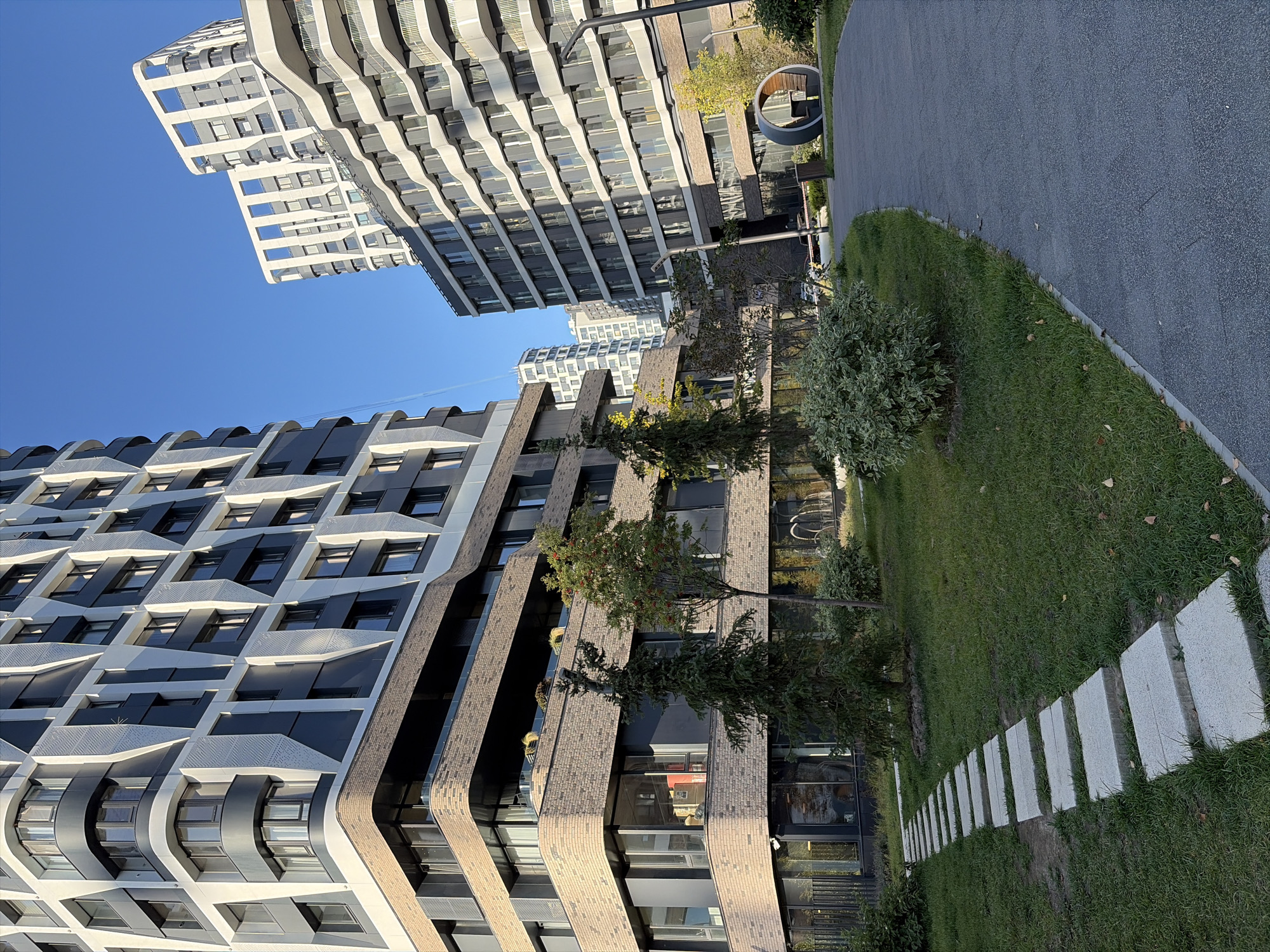 The image depicts a modern residential complex in Moscow, Russia, characterized by its distinctive architectural design. Tall buildings with striking facades, featuring a combination of dark gray, white, and brick elements, dominate the scene. The buildings have numerous windows and balconies, with some showcasing curved architectural features.
In the foreground, a well-maintained courtyard area is visible. A pathway made of square stone slabs winds through a lush green lawn dotted with small trees and shrubs. To the right, a paved walkway curves around the lawn. Further back, a unique circular seating area with a table is a prominent feature. The sky above is a clear, bright blue, indicating a sunny day. The overall impression is of a contemporary urban living space designed with an emphasis on aesthetics and greenery.