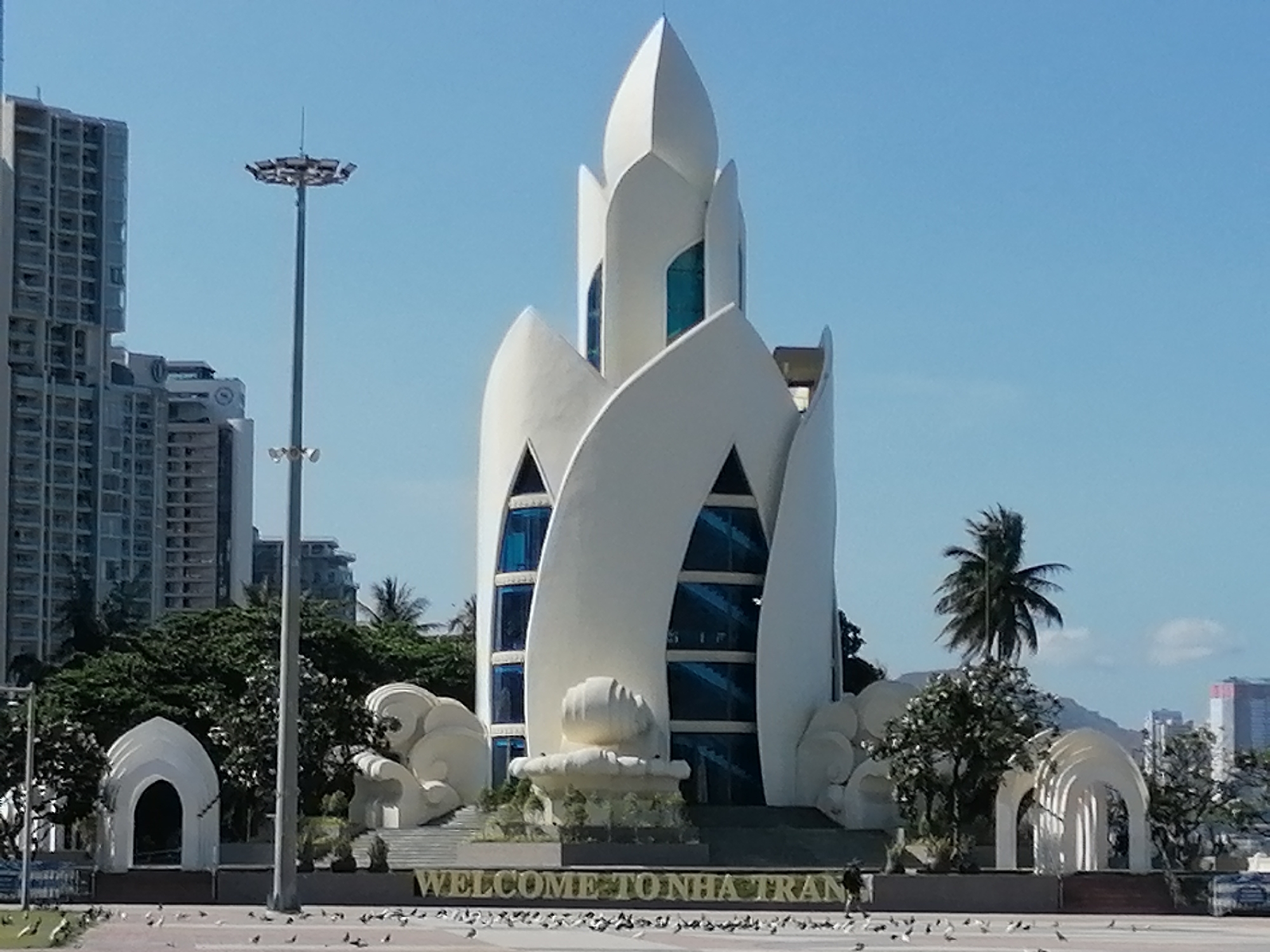 The image shows the Lotus Tower Welcome Center in Nha Trang Ward, Vietnam. The tower is white, stylized as a lotus flower, with blue-tinted windows. In front of the tower is a fountain and the words "WELCOME TO NHA TRANG" in gold lettering. To the left and right of the tower are white arched structures and sculptures. A tall light pole stands on the left. In the background are buildings, trees, and a clear blue sky. Many pigeons are scattered on the ground in front of the welcome center. A palm tree is seen near the tower.