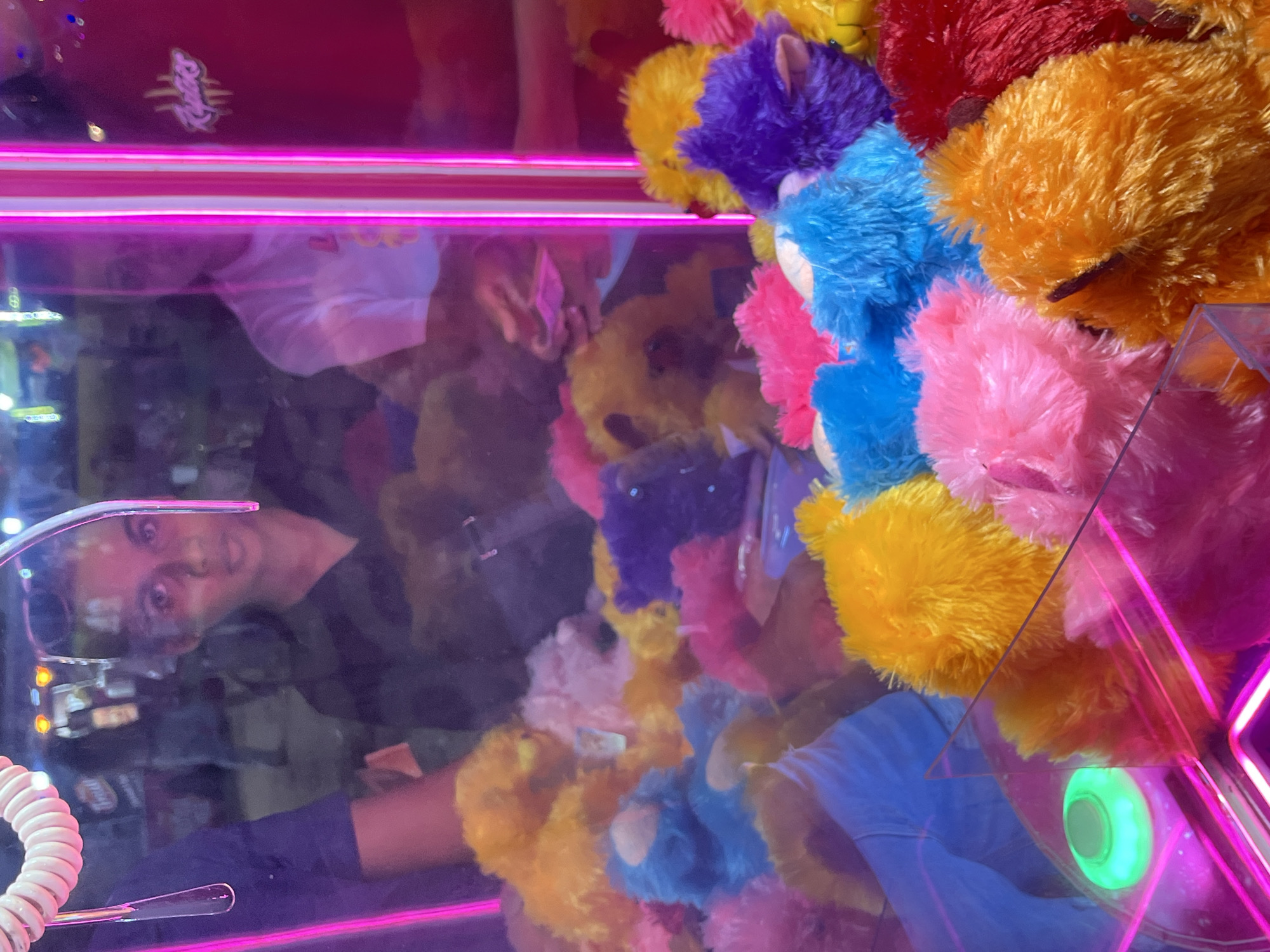 The image shows a person reflected in the glass of a claw machine filled with colorful, fuzzy stuffed animals. The stuffed animals are various bright colors, including yellow, orange, pink, blue, and purple, and appear to be small, round, and bear-like. They are densely packed inside the machine, making it difficult to discern individual toys clearly. The machine has bright pink neon lights illuminating the top and sides. The reflection shows a person wearing a dark shirt, their face tilted and looking towards the viewer. The lighting in the machine is dim, with the neon lights being the most prominent light sources. The reflection of the person suggests a casual, indoor setting, possibly an arcade or amusement area. The overall impression is of a vibrant and playful environment, with the stuffed animals serving as the main objects of interest and the reflection adding a human element to the scene. The location cue of "Habaraduwa, Sri Lanka" does not provide any direct visual information in the image itself but suggests a tropical or subtropical setting where such entertainment venues might be found. There is text visible on a pink tag attached to one of the stuffed animals, which appears to be a logo or brand name, but it is too blurry to read.