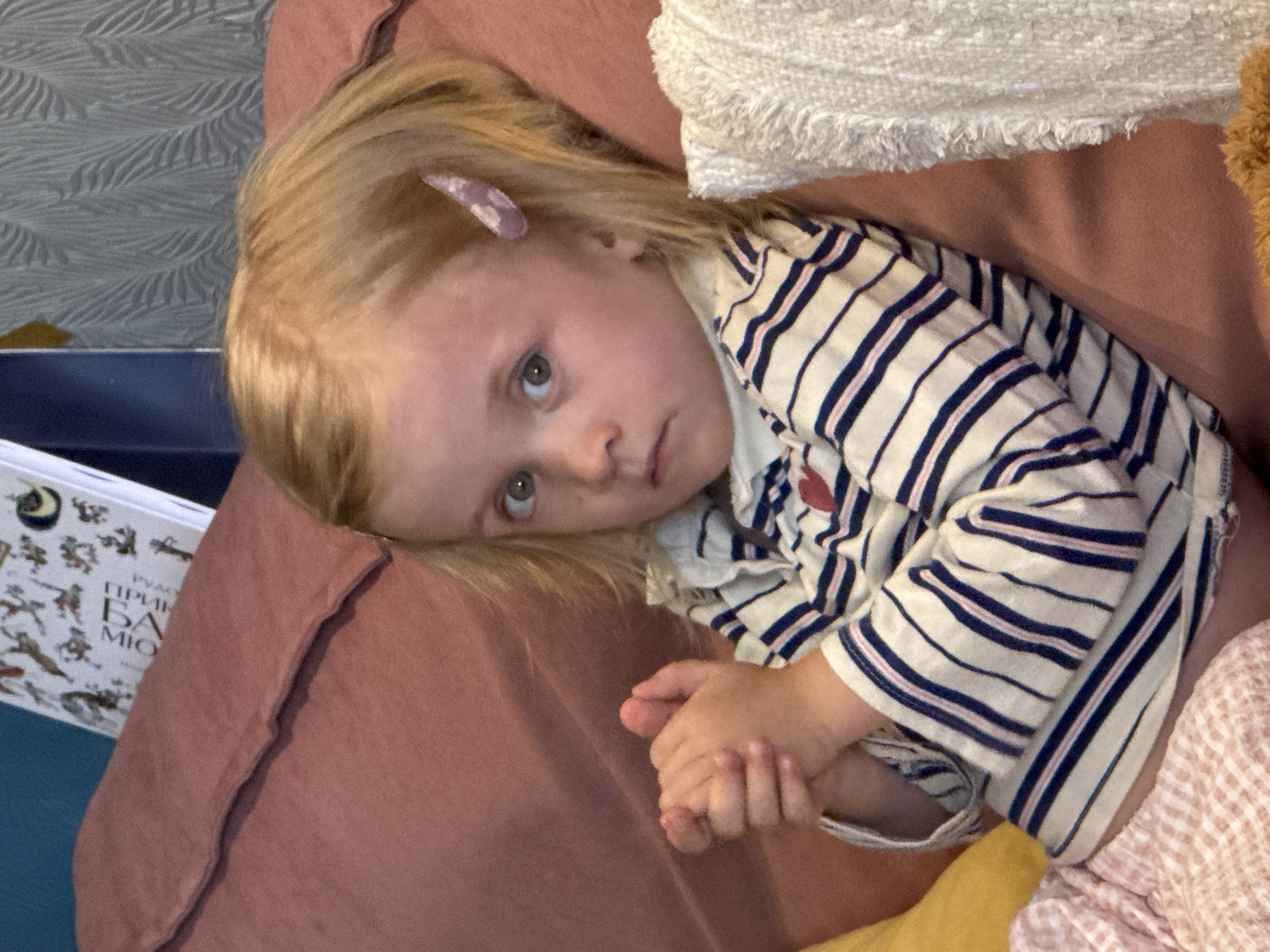 A young, fair-skinned child with blonde hair is lying on a reddish-brown pillow or cushion. The child has a light purple clip in their hair. They are wearing a long-sleeved striped shirt with navy and pink stripes on a white background. The child is looking upwards with a somewhat thoughtful expression. Their hands are clasped together. Part of a book with Cyrillic text is visible in the lower left corner. A white textured fabric is visible at the top right. There is no definitive evidence or visual cues in the provided image to pinpoint the location in Moscow, Russia, or any other specific location.
