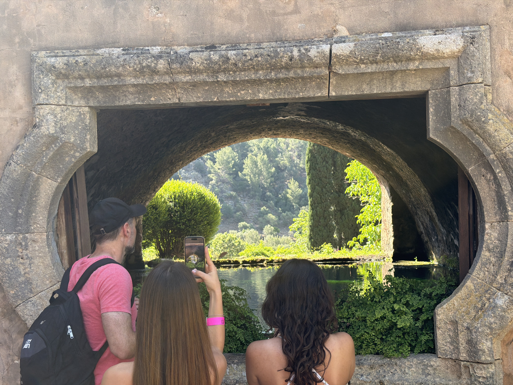 Three individuals stand before a stone structure featuring a curved archway, framing a tranquil scene of water, greenery, and distant hills in Bunyola, Spain. A man in a pink shirt and black cap stands to the left, wearing a black backpack. A woman with long, straight hair holds up a cell phone, presumably taking a photograph of the view. Another woman with curly hair is positioned to the right, with her back to the camera. The setting appears to be a garden or park-like area, with abundant vegetation. The stone archway is likely part of a historical structure or water feature.