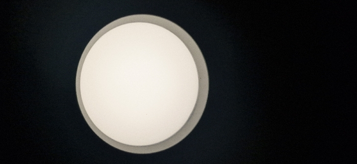 A close-up shot features a circular ceiling light against a dark background. The light has a distinct circular frame with a slightly rounded inner surface, suggesting a diffused glow. The color of the light is warm white or off-white, in stark contrast to the surrounding darkness. The image is simple, emphasizing the round light fixture and its gentle illumination.