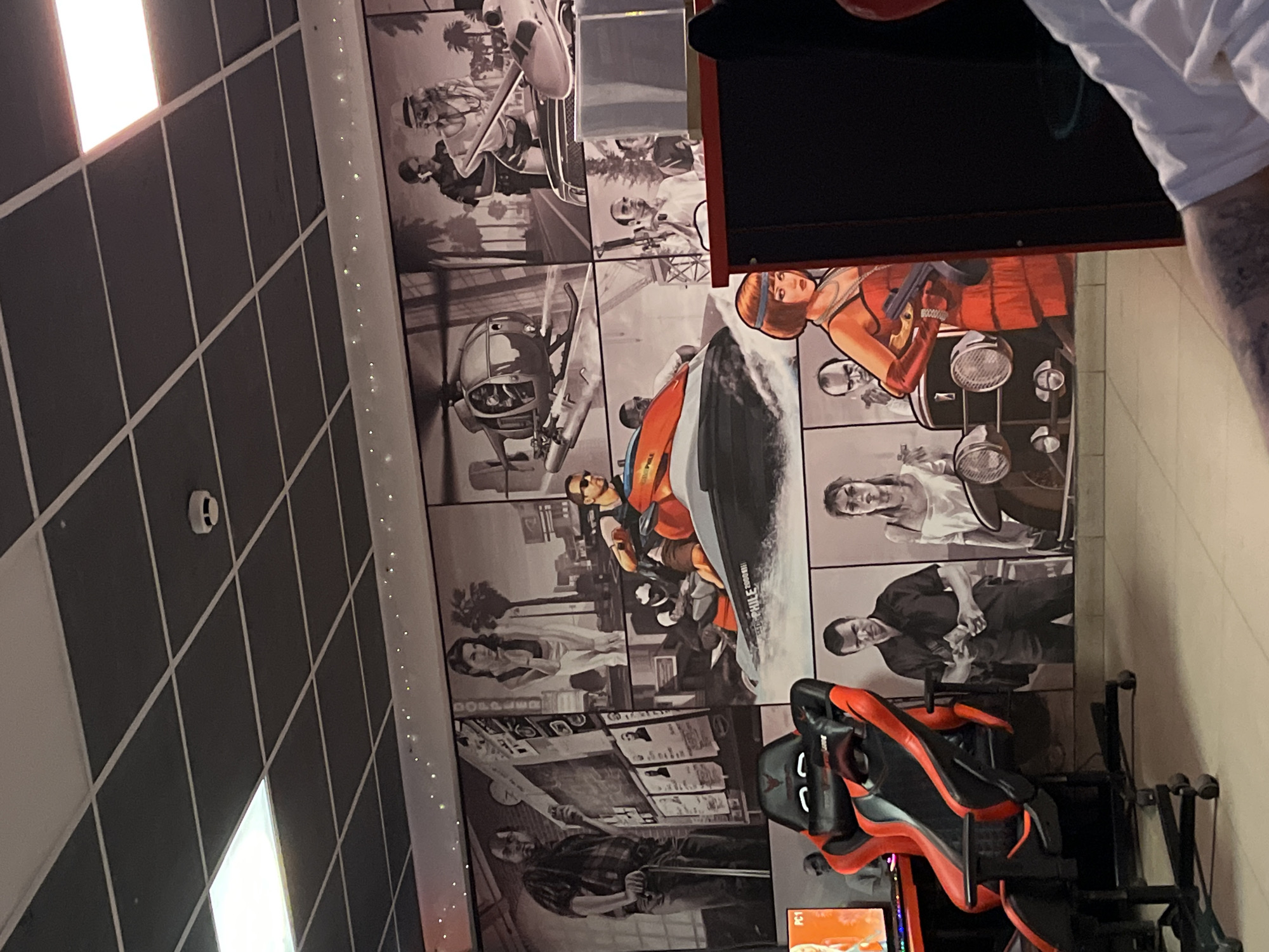 The image depicts a wall mural in black and white, featuring scenes that appear to be inspired by the Grand Theft Auto video game series. The mural is divided into multiple panels showcasing various characters and vehicles. In the lower left corner, a gaming chair with red and black accents is visible. To the left of the chair, a computer monitor displays "PC1". The mural includes a man standing with a scooter, a helicopter, a jet ski with riders, several figures in different poses, and a woman in a red dress holding a firearm next to a vintage car. The environment appears to be indoors, possibly a gaming cafe or a personal entertainment space, with a tiled ceiling and fluorescent lighting. The location is stated to be Chaikovsky, Russia, though this is not visually evident from the image itself.