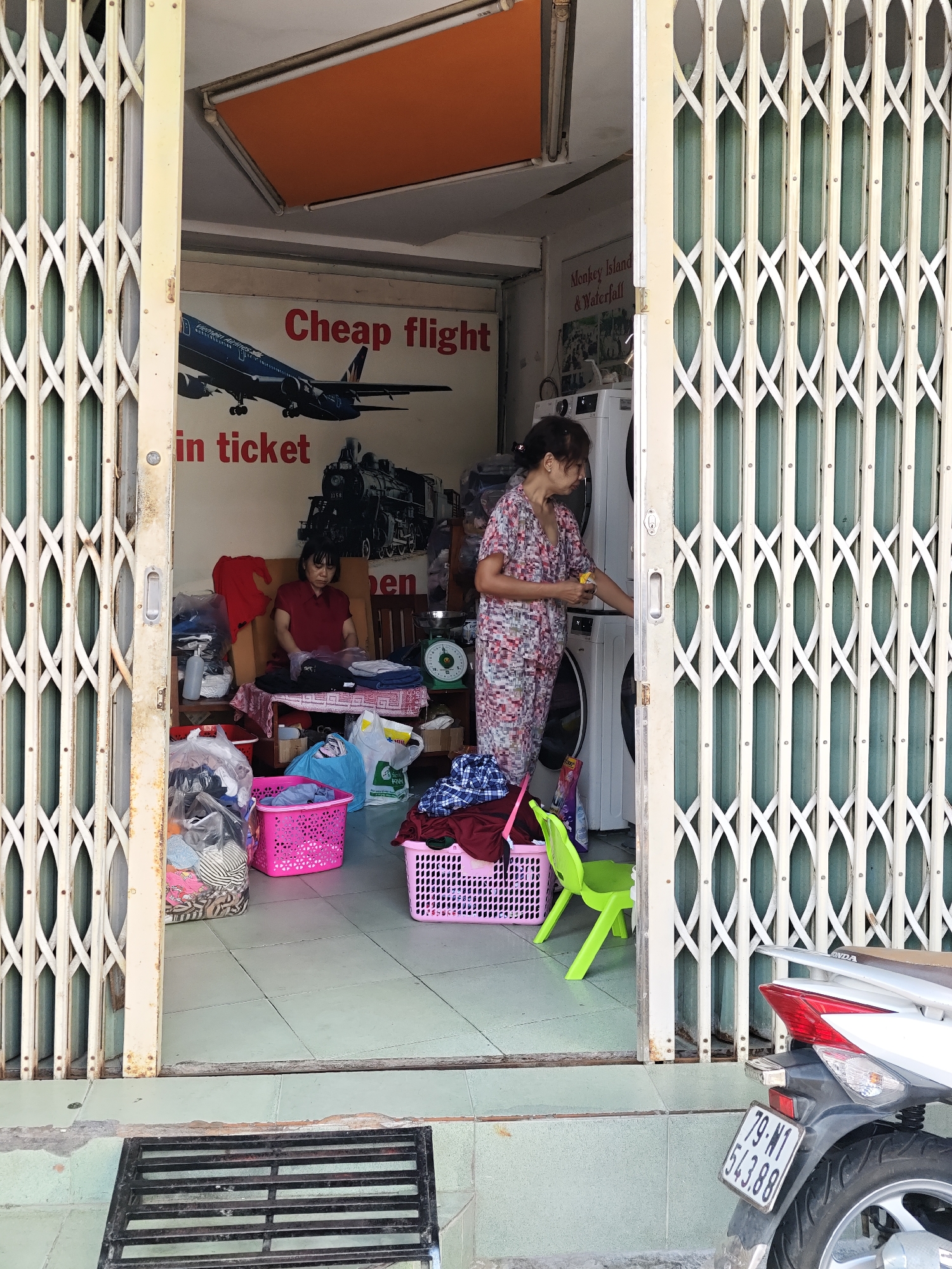The image shows the interior of a shop with its metal gate partially open. The shop appears to be a laundromat, with several washing machines visible in the background. A woman in a floral-patterned outfit is standing next to a washing machine, holding a bottle and possibly dispensing detergent. Another woman, wearing a red shirt, is seated behind a table where laundry is being folded. There are piles of laundry in baskets and bags throughout the shop. A poster on the wall advertises "Cheap flight in ticket". A weighing scale is visible on a counter. Outside the shop, a white motorcycle is parked, with a license plate that reads "79-N1 543.88". The overall environment suggests a commercial establishment in a city. The license plate number indicates the location is likely within Vietnam, specifically Nha Trang Ward, given the "79" prefix for the province of Khánh Hòa, where Nha Trang is located.
