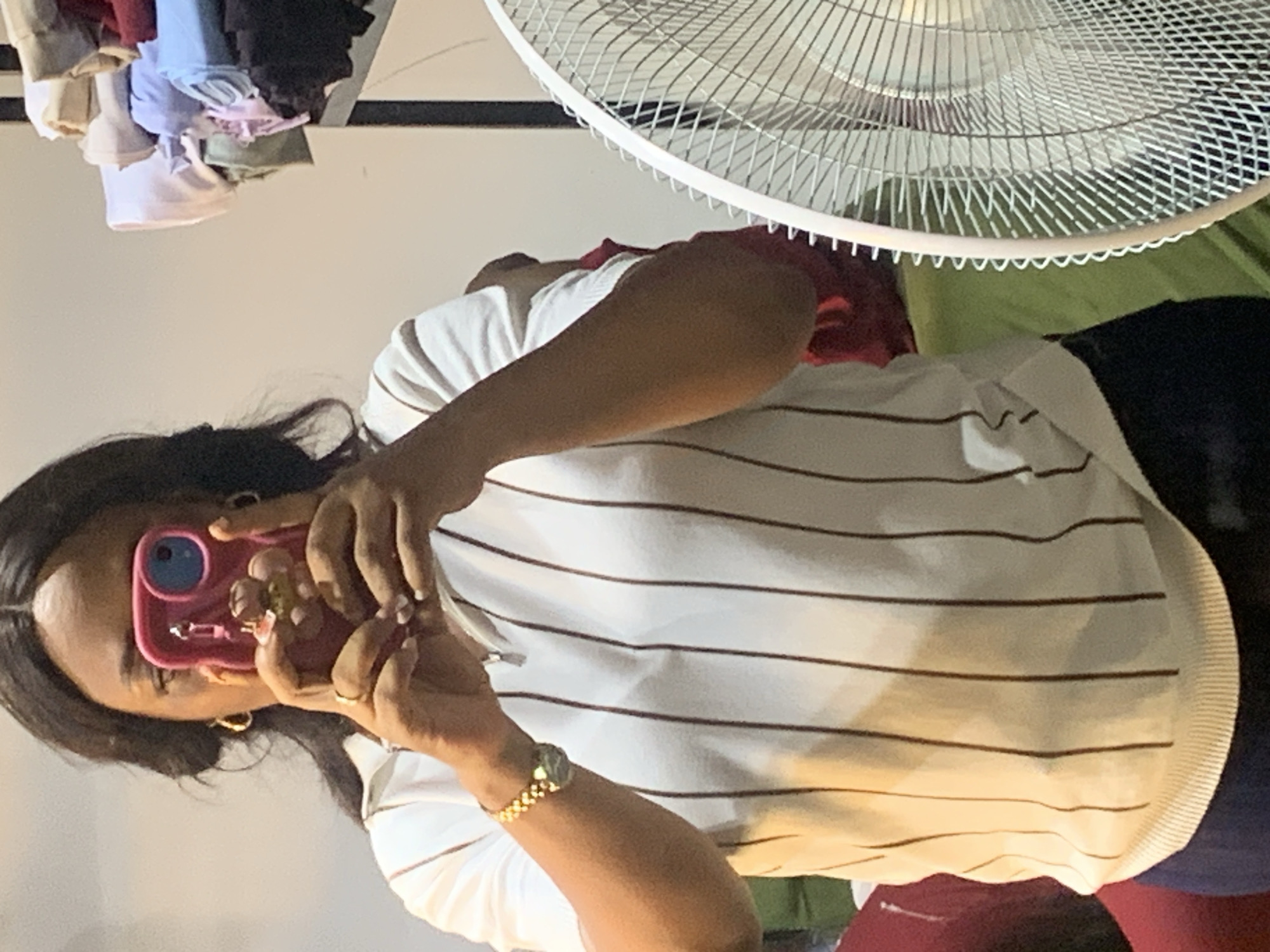 A person is taking a selfie indoors. The person is wearing a striped shirt and has a gold watch on their left wrist. A white electric fan is partially visible in the upper right corner. Clothing items hang in the upper left. The location is Narayi, Nigeria.