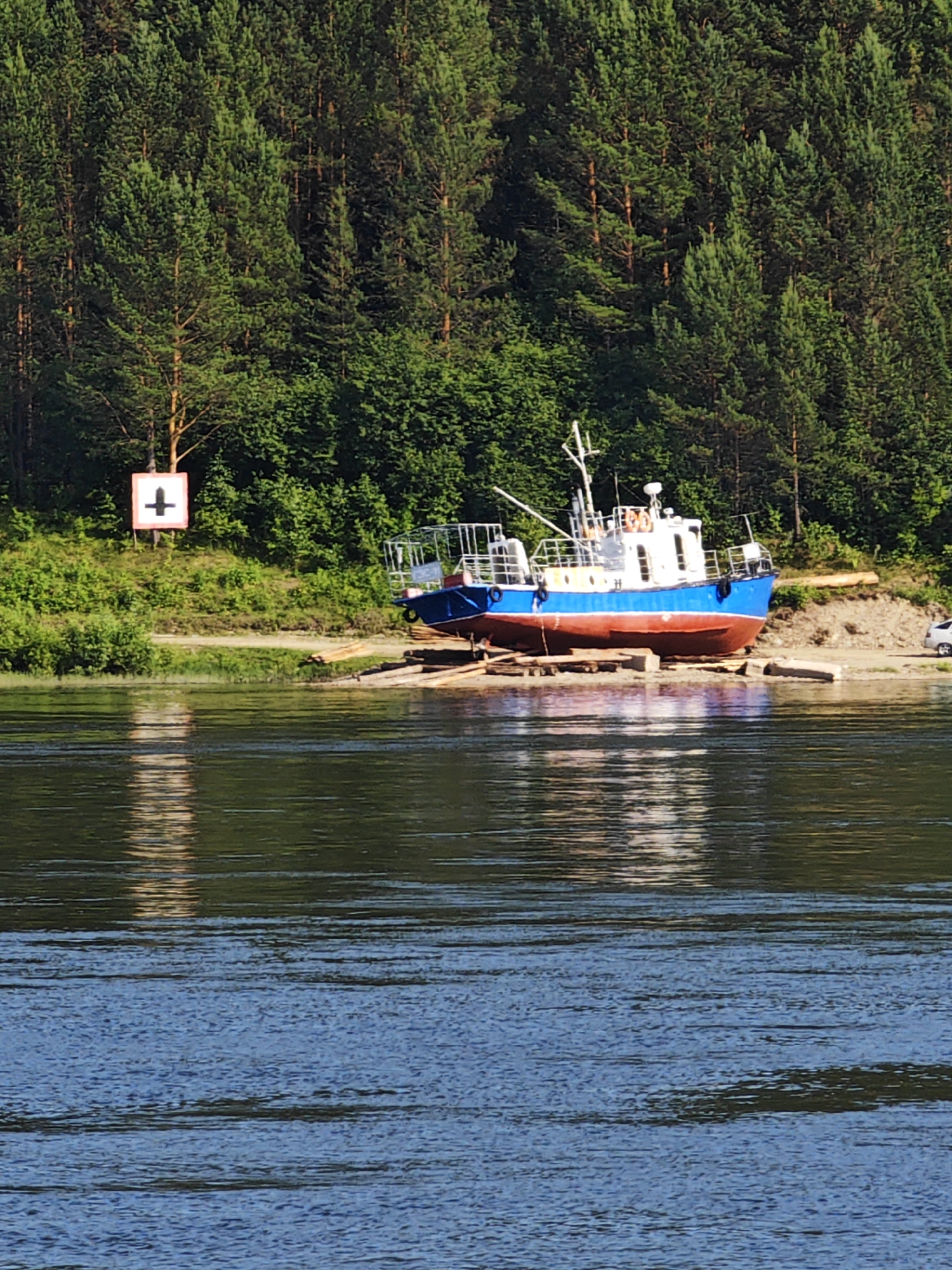 A blue and red vessel is beached on the shore of the Chulym River, adjacent to the city of Predivinsk, Russia. The vessel rests on a wooden support structure. A green forest borders the riverbank. A white and red sign with a black cross symbol is visible on the bank. The water reflects the surrounding trees.