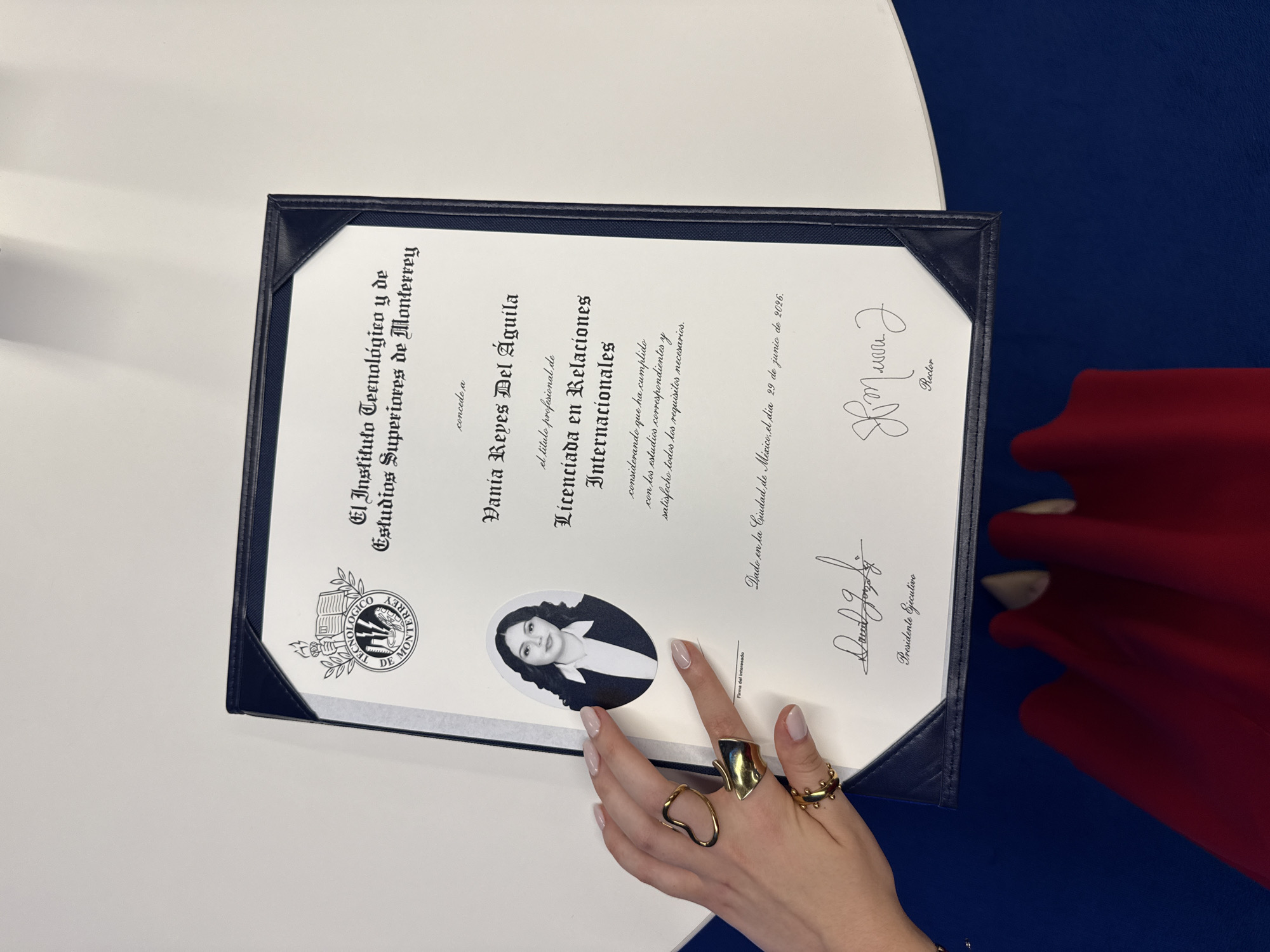The image displays a framed diploma from "El Instituto Tecnológico y de Estudios Superiores de Monterrey," awarded to Vania Reyes Del Águila for a degree in International Relations. A black and white portrait photo is included on the diploma. The diploma is signed by the President and Rector and specifies that the degree was conferred in Mexico City on June 29, 2026. A hand wearing multiple gold rings is partially visible, holding the diploma. The background consists of a white surface and a portion of blue and red material.