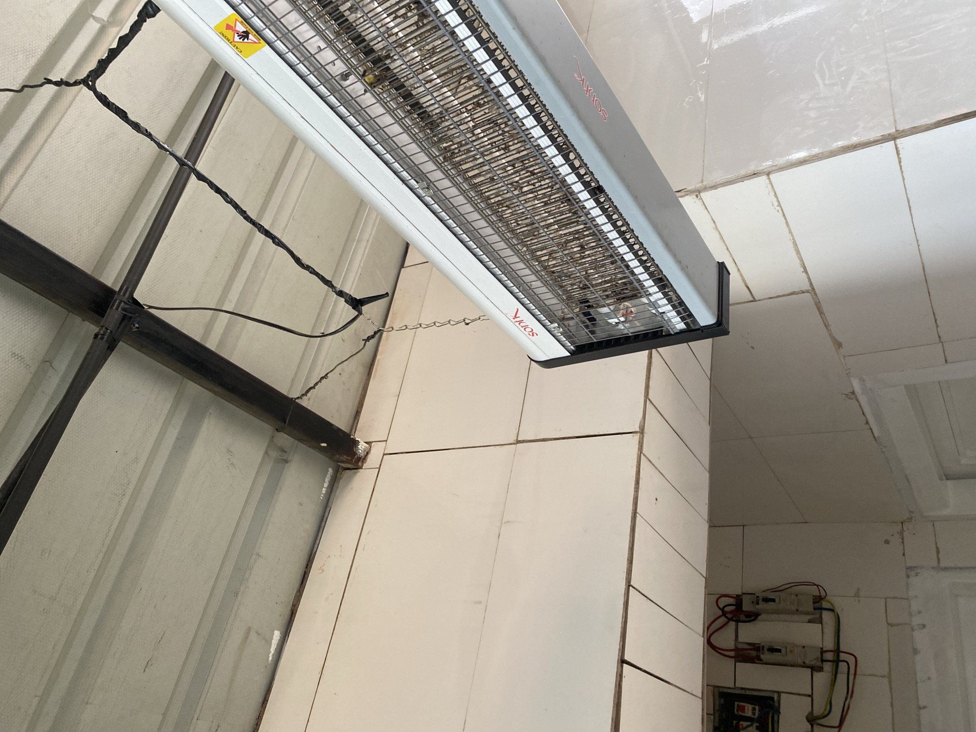 The image depicts an interior space, location Unknown, Unknown, characterized by utilitarian construction. The ceiling consists of light grey or off-white corrugated metal panels supported by dark metal beams. Black electrical wiring, secured with black tape, is visibly routed along these beams.
Suspended from the ceiling by a metal chain and an electrical cord wrapped in black tape is a large, rectangular electronic insect killer, branded "soniK" in red lettering. The device's interior grid is densely packed with a significant accumulation of dead insects, indicating extensive use and a lack of recent maintenance. A yellow triangular warning sticker with a "CAUTION" text and a hand symbol is affixed to the side of the unit.
The walls are covered with white or off-white rectangular ceramic tiles, exhibiting visible grout lines that appear aged or discolored in some areas. In the lower-left quadrant of the image, an electrical installation is visible on the tiled wall. This includes an open electrical box and two wall-mounted circuit breakers or switches, with red, black, and yellow wires connected and partially exposed. The lower right section of the image shows a portion of a white-framed doorway or opening.