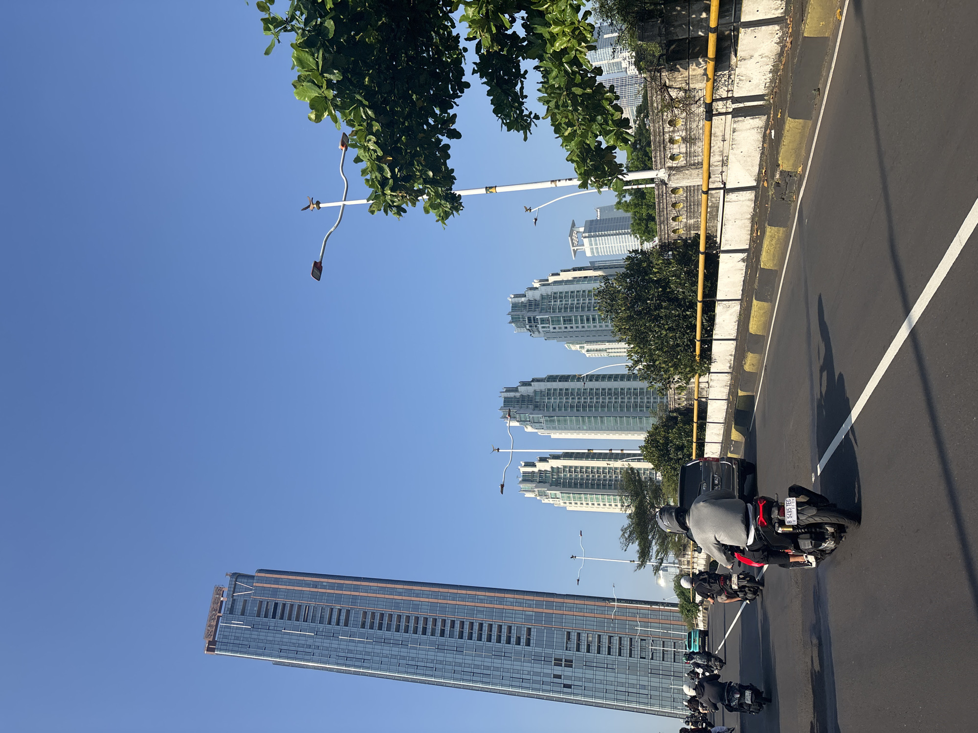 The image captures a street scene in Central Jakarta, Indonesia, notable for its blend of urban and natural elements. A line of motorcycles travels along a gray asphalt road with a solid white line marking the lane. The riders, clad in helmets and casual attire, create a procession. Dominating the skyline are modern buildings, including several high-rise apartment buildings and a tall glass skyscraper. Trees with dense green foliage frame the upper part of the view. The sky is clear and blue. The street is lined with a yellow and gray concrete barrier, adding to the urban landscape. The presence of distinctive lighting fixtures on curved poles is a unique feature of the urban environment. The scene is captured in daylight with strong sunlight.