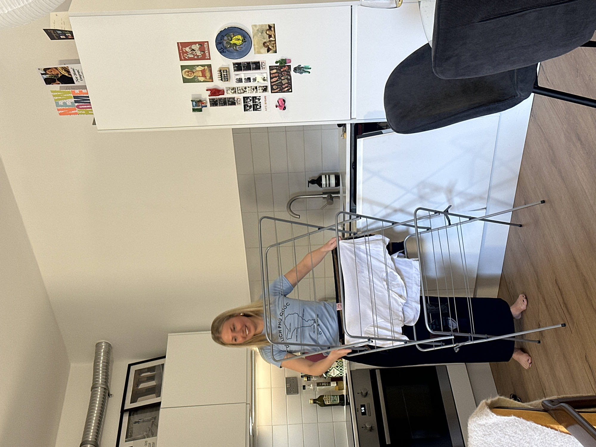 A woman with blonde hair is putting white laundry on a silver drying rack in a modern kitchen. The kitchen has white cabinets, a stainless steel oven, and a tile backsplash. There are magnets on the refrigerator, and framed pictures on the wall. The location appears to be a residential kitchen in London, United Kingdom.