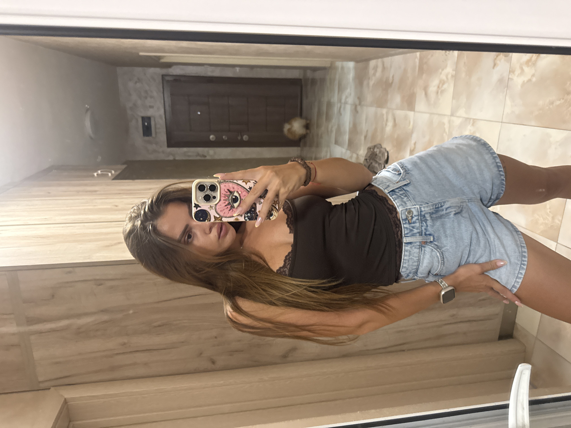 A young woman with long brown hair takes a mirror selfie in what appears to be a home hallway. She is wearing a dark brown camisole with lace trim and light blue denim shorts. She is also wearing a silver smartwatch on her left wrist and several bracelets on her right wrist. Her phone, with a pink case featuring an eye and stars design, is held up to take the photo. In the background, a door is visible at the end of the hallway, and a small, fluffy dog with light brown and white fur is walking away from the camera. The hallway has tiled flooring and wooden wardrobe doors on the left. The overall scene suggests a casual, domestic setting. The location is not specified in the image or its metadata, therefore it cannot be definitively identified as Plovdiv, Bulgaria.