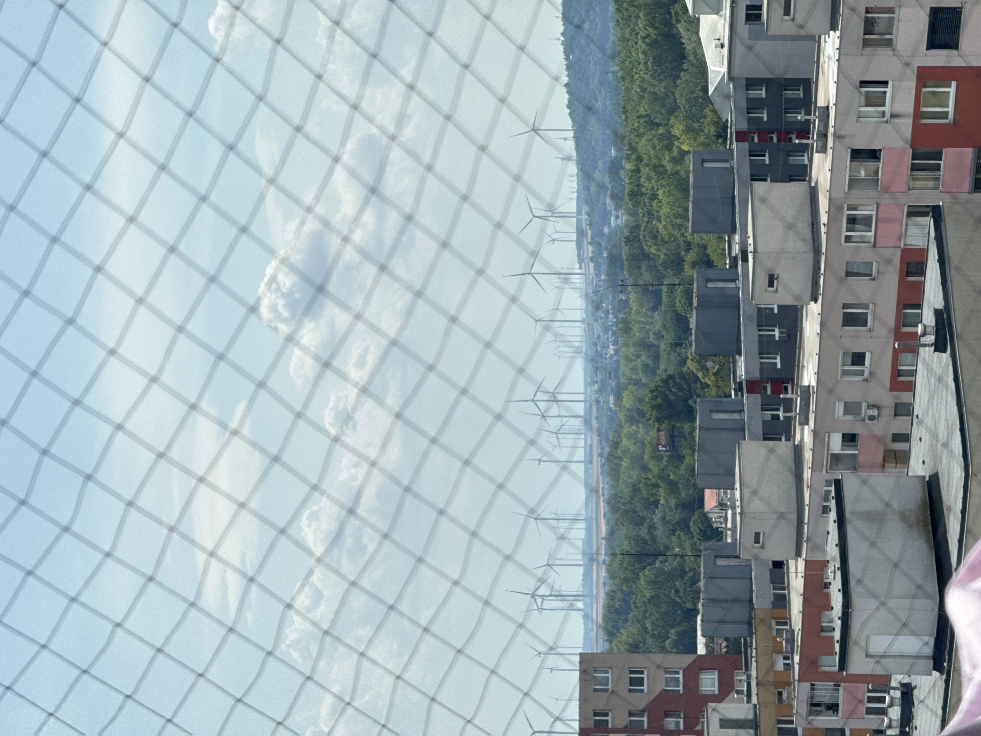 The image shows a view of a city with numerous wind turbines in the background, seen through a diamond-patterned net. In the foreground and midground, there are several multi-story residential buildings, some appearing to be Soviet-era apartment blocks. The buildings have various colors, including beige, red, and grey, and feature numerous windows. Some buildings have flat roofs with visible ventilation systems or small structures on top. The sky is blue with scattered white clouds. The overall scene suggests an urban or suburban environment on a clear day, with a focus on residential architecture and renewable energy infrastructure. The location is identified as Bratislava, Slovakia, likely a view from a higher vantage point overlooking the city.