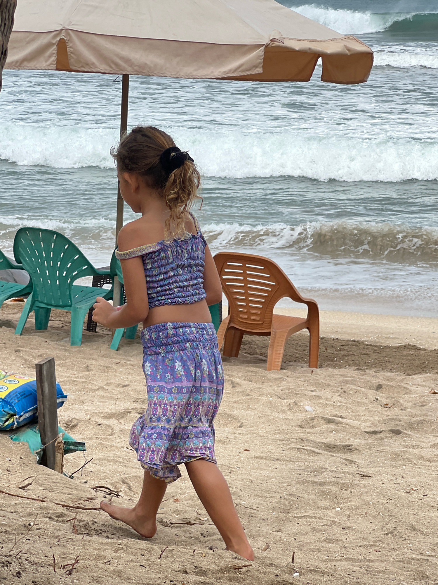 In Kuta, Indonesia, a young girl with blonde curly hair tied back with a black scrunchie walks on a sandy beach. She is wearing a purple off-the-shoulder cropped top and matching shorts with a floral pattern. Her left leg is extended forward, mid-stride. In the background, the ocean waves crash onto the shore under a cloudy sky. Several beach chairs, two green and one orange plastic chair, are scattered on the sand. A large beige beach umbrella is partially visible in the upper portion of the image, providing shade. A wooden post with a blue and yellow bag is situated to the left of the girl. The scene captures a casual moment at the beach.
