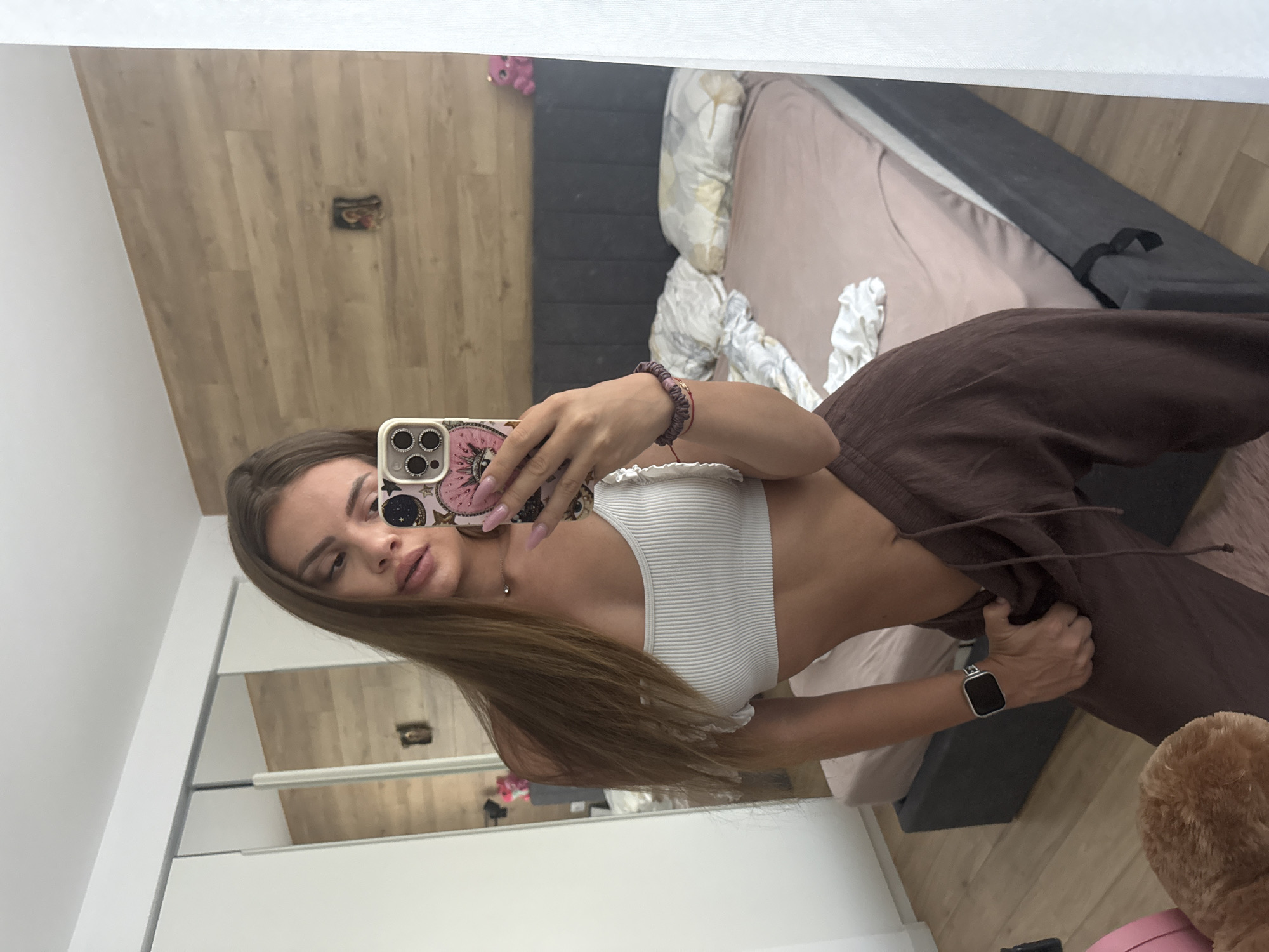 The image depicts a mirror selfie of a young adult female taken inside what appears to be a modern bedroom in Plovdiv, Bulgaria. The female subject, positioned centrally, has long, straight brown hair extending to her chest. She is wearing a white ribbed short-sleeved crop top and loose-fitting, dark brown drawstring pants, which she is pulling slightly with her left hand. Her right hand holds a light-colored iPhone with a distinctive multi-lens camera array and a pink, patterned case featuring an "evil eye" motif. She has pink nail polish on her long fingernails and wears a thin, silver-colored necklace, multiple bracelets on her right wrist (including a dark purple braided one), and a silver-toned smartwatch on her left wrist.
The background, a reflection in the mirror, shows a room with a light wooden plank accent wall behind a dark gray upholstered bed frame. The bed is made with a light peach/pink fitted sheet and has several pillows, one with a yellow and gray geometric pattern, and some rumpled white bedding. Above the bed, on the wooden wall, hangs a small framed religious icon. To the left, a white sliding door wardrobe or closet is visible, reflecting a portion of the room. A small pink plastic object, possibly a toy bear, is visible on a surface above the bed in the reflection. The floor is light wood, and a fuzzy pink rug is partially visible in the lower-center foreground of the reflection. The lighting is bright, suggesting natural light from a window or well-lit interior.