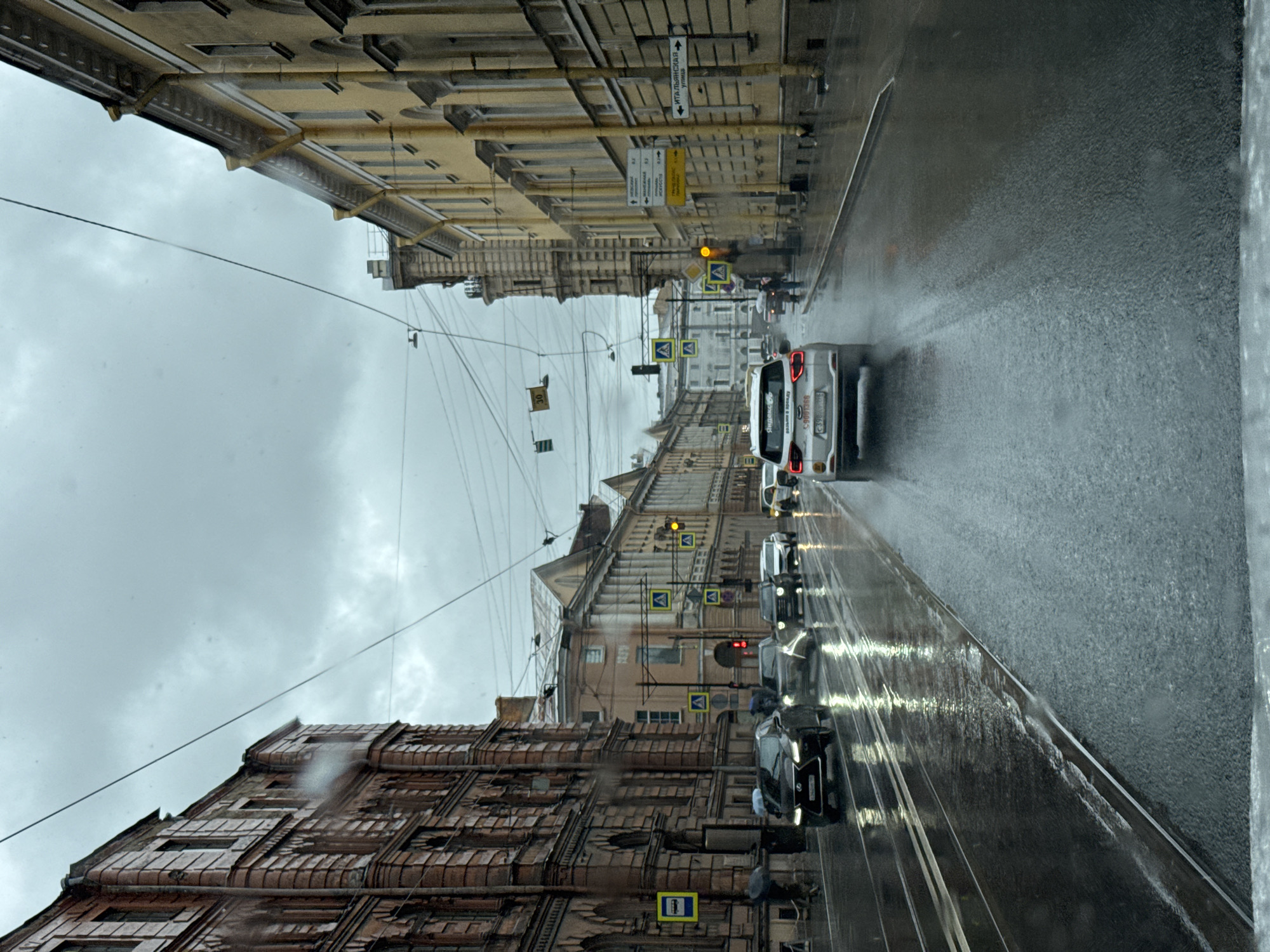 The image depicts a rainy day scene on a street in Saint Petersburg, Russia, likely in the historic center given the architecture. The street is wet and reflects the lights of passing vehicles. Several cars are visible, including a white SUV in the foreground, a dark sedan and other vehicles further down the road. Pedestrians with umbrellas can be seen on the sidewalks. The buildings lining the street are multi-story structures with classic architectural details, consistent with the style found in Saint Petersburg. Overhead, utility wires crisscross the sky, and traffic lights and street signs are present. The sky is overcast and grey, indicating inclement weather.