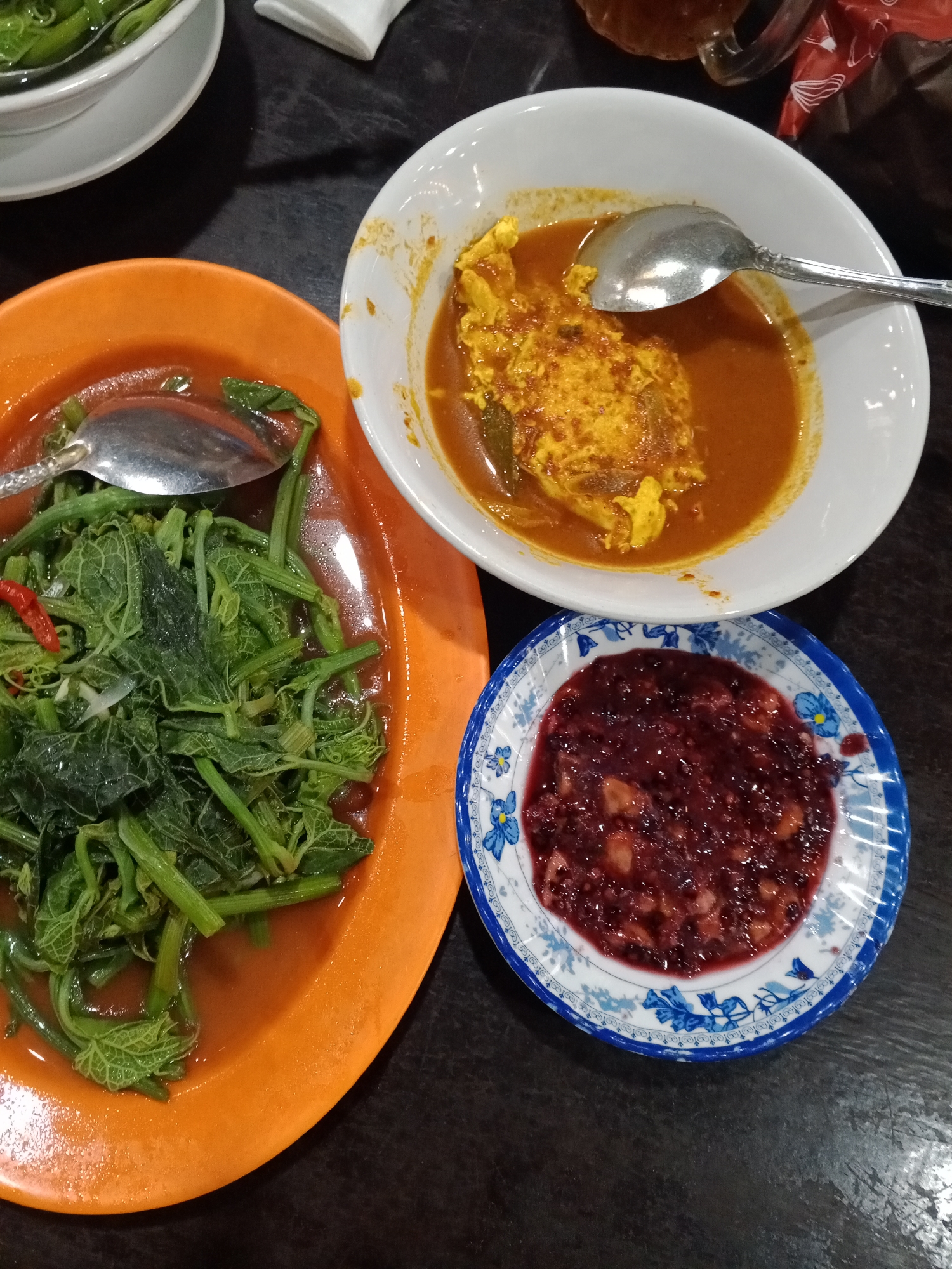 Three plates of food are arranged on a dark, possibly wooden, table. In the foreground, an orange plate holds a serving of green leafy vegetables in a light brown sauce, with a single red chili pepper visible among the leaves. A silver spoon rests on the edge of this plate. To the right of the orange plate, a small blue and white patterned plate contains a dark red, chunky condiment, possibly sambal. Behind these plates, a white bowl contains a yellow and orange dish, appearing to be a stew or curry with pieces of egg. A silver spoon is partially submerged in this dish. In the upper left corner, a white bowl, partially visible, contains green food, possibly more vegetables. The lighting suggests an indoor setting, likely a restaurant. The overall scene depicts a meal.