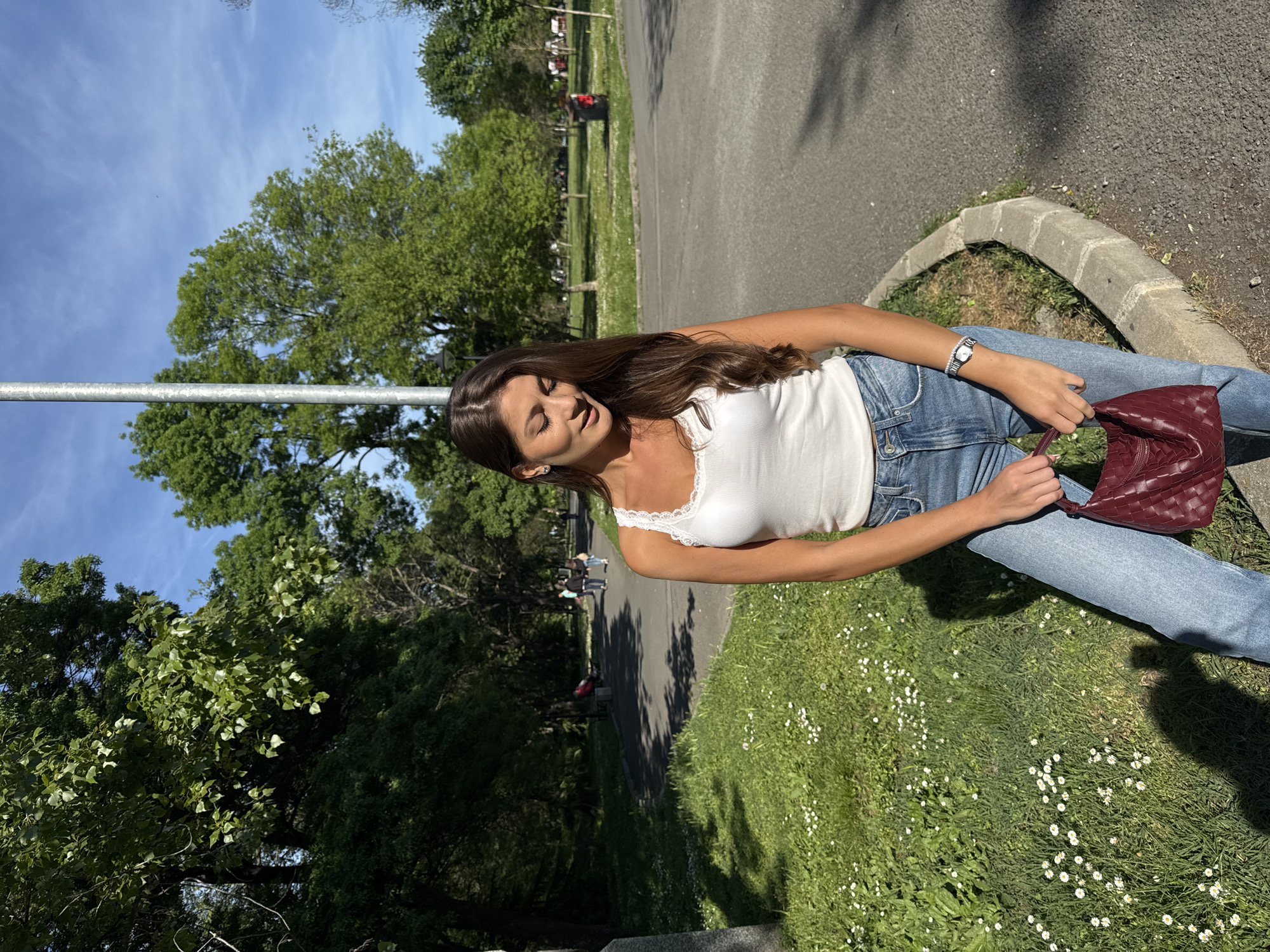 The image shows a young woman in a park-like setting. She is wearing a white tank top, blue jeans, and a watch. She is holding a burgundy woven handbag. The background includes trees, grass, and a paved path. The location cannot be determined from this image.