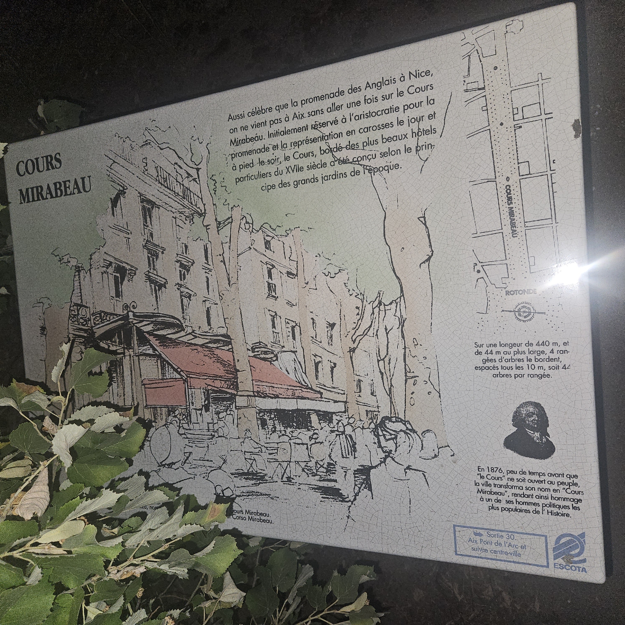The image shows a historical information plaque in the city of Rousset, France. The plaque features an artistic depiction of a street scene, likely the Cours Mirabeau, with buildings and people. Text on the plaque describes the Cours Mirabeau, its history, and its length and width. There is also a map of the area. The plaque is partially obscured by foliage. The text is in French. The overall impression is that of a historical marker providing context and information about a significant location within the city.