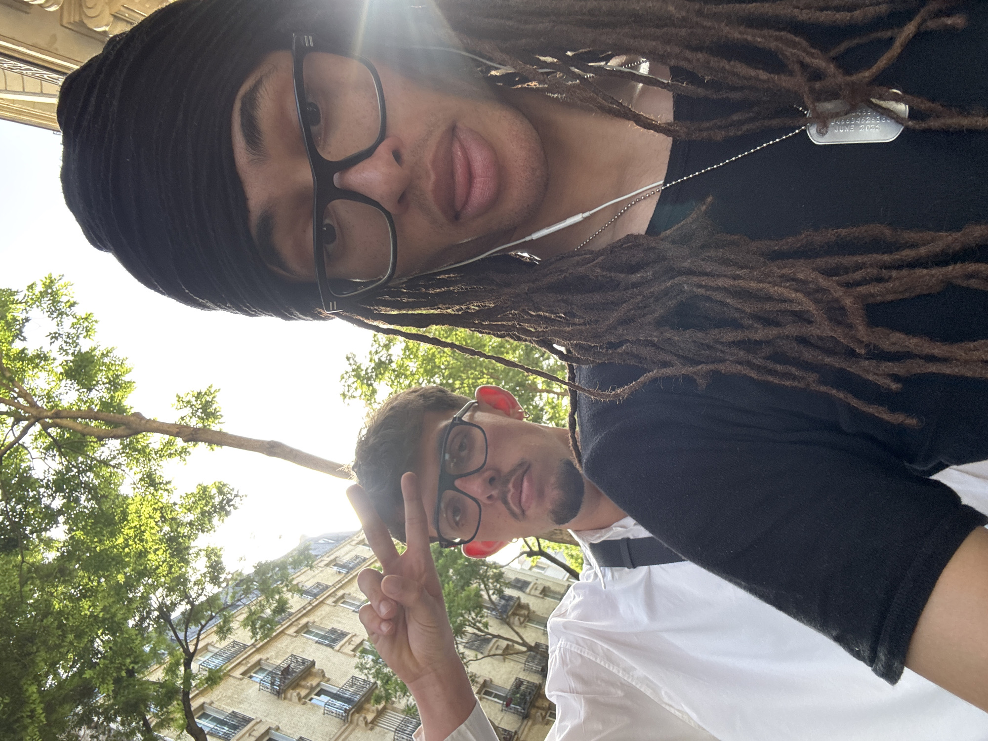 The image shows two young men taking a selfie outdoors. The man at the top wears a black beanie, glasses, and has dreadlocks. He wears a black shirt and a dog tag necklace. The second man is smiling and making the peace sign. He wears glasses and a white shirt. In the background, there are trees and a building with balconies, typical of architecture in Paris, France. Sunlight is streaming into the frame, suggesting the photo was taken during the day.
