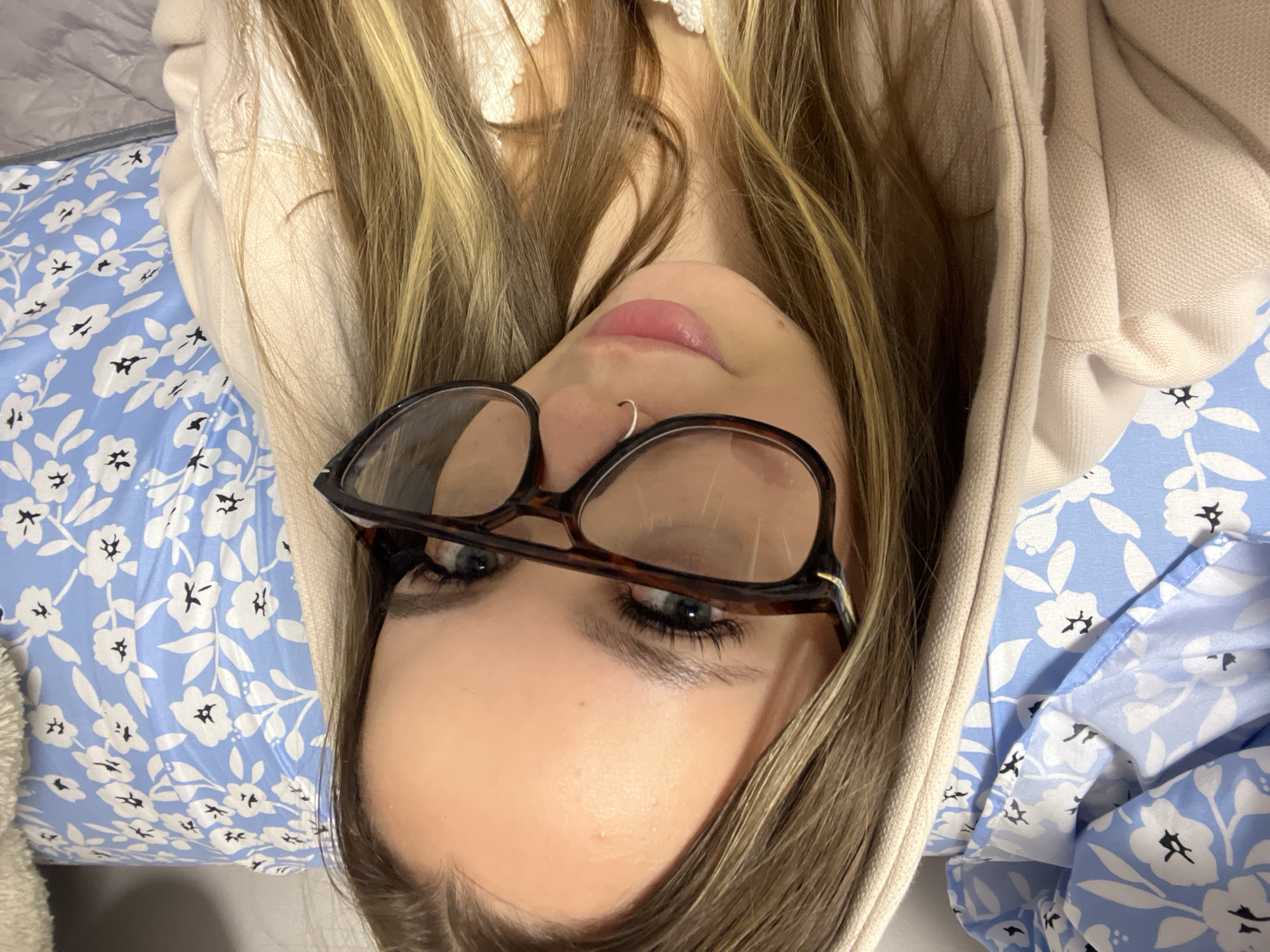 An image captures a young individual, likely a female, with long, blonde-streaked brown hair, lying down and looking towards the camera. The person wears dark-rimmed, possibly tortoise-shell, glasses pushed down on their nose, revealing light blue or grey eyes with visible eyelashes and defined eyebrows. A small silver hoop nose ring adorns their left nostril, and their lips are full with a glossy, pink finish.
The subject is wearing a light beige or cream-colored hooded garment, possibly a hoodie or sweatshirt, with its hood partially obscuring the sides of their head. Beneath or around them is a vivid blue fabric adorned with a repeating pattern of small white flowers, each with a dark center, and white leaves. This fabric appears to be either clothing worn underneath the hoodie or bedding, suggesting an indoor setting. The image is oriented such that the subject's head is on the left side, indicating they are lying on their back or side.
The overall impression is casual and relaxed, with soft lighting illuminating the scene. No specific activity is depicted beyond the pose itself. The location context for this scene is Krasnodar, Russia. There is no visible text in the image.
