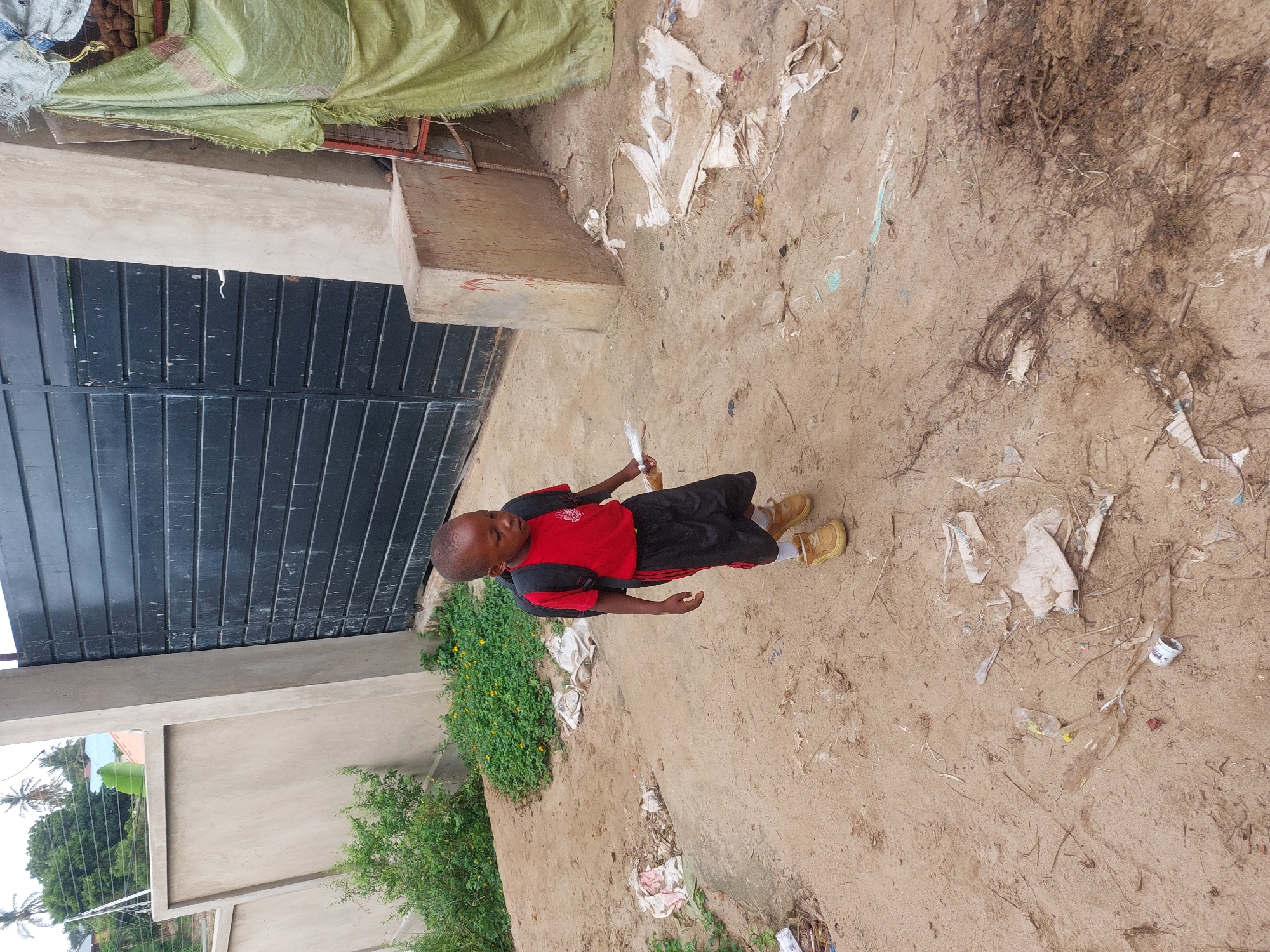 The image depicts a child in a red shirt, black shorts, and tan shoes standing on a dirt ground with scattered debris. A gate and part of a building or wall are visible to the left, alongside some greenery. The environment appears to be a residential or semi-urban area, with trash and loose material strewn on the ground. Without more contextual clues, it's impossible to verify that the location is specifically in Dar es-Salaam, Tanzania.