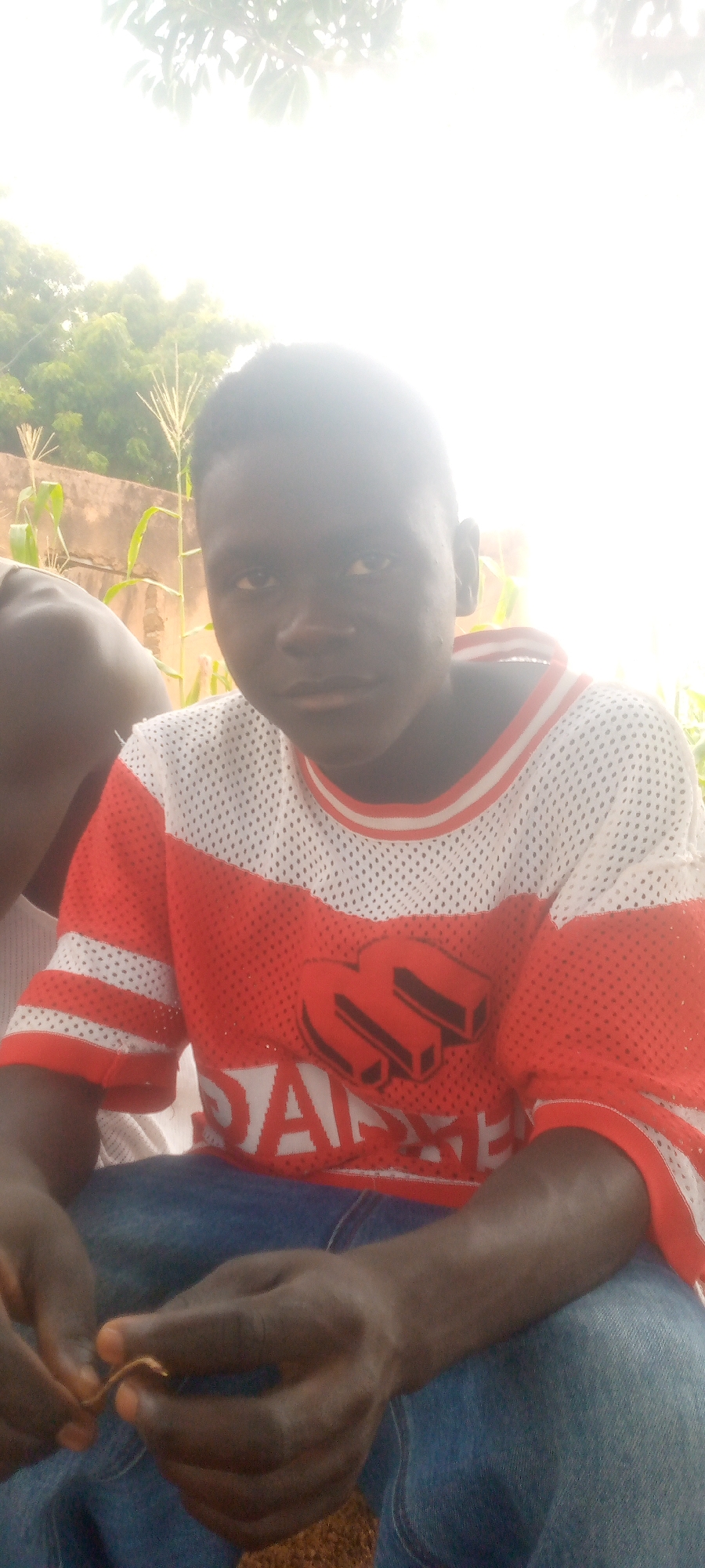 A young boy is pictured outdoors, sitting in what appears to be a rural or semi-rural setting. He is wearing a red and white mesh t-shirt with white accents on the sleeves and collar. His t-shirt has a large, black, three-dimensional graphic on the front, which is partially obscured. He is also wearing blue jeans. His hands are clasped in front of him, and he appears to be holding a small, thin, brown object. The background shows a wall, some corn stalks, and greenery, suggesting an agricultural environment. The lighting is bright, with some overexposure in the upper portion of the image, indicating natural sunlight. The boy's face is turned slightly towards the camera, and he has a neutral expression. The overall scene is casual and appears to be a candid shot. The location is not specified in the prompt, hence 'Unknown, Unknown'.