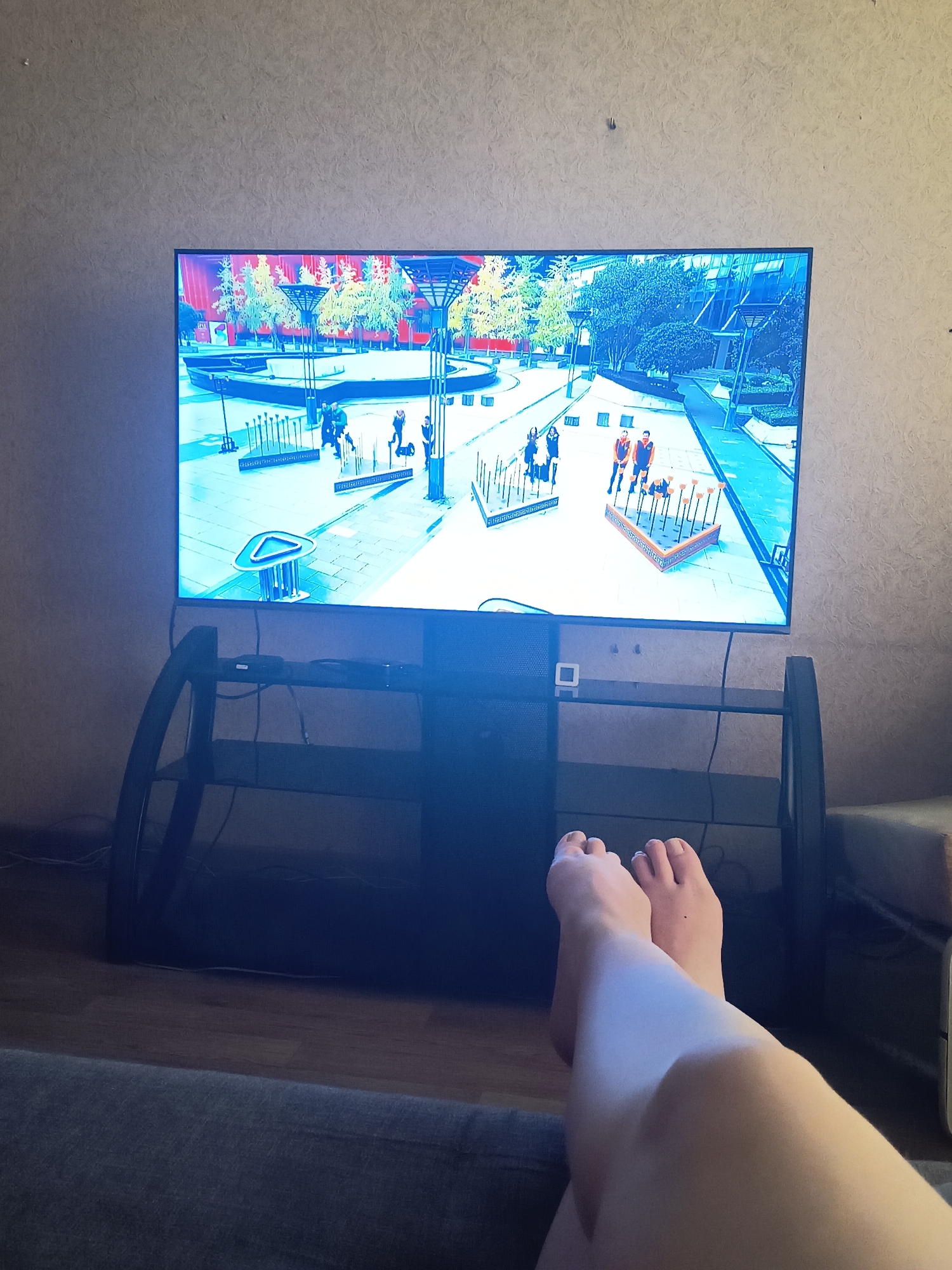 The image shows a large flat-screen television displaying a video game. The game appears to be set in an outdoor urban environment with stylized trees and modern architecture in the background. Several figures are visible on the screen, engaged in various activities. In the foreground of the image, a person's legs and feet are visible, relaxed as they view the television. The room is dimly lit, suggesting it might be evening or nighttime. The television is placed on a black media stand, with cables visible. The wall behind the television has a textured wallpaper. The overall scene conveys a sense of leisure and entertainment, with the person enjoying a video game. The location context is Blagoveshchensk, Russia. There is no visible text in the image.