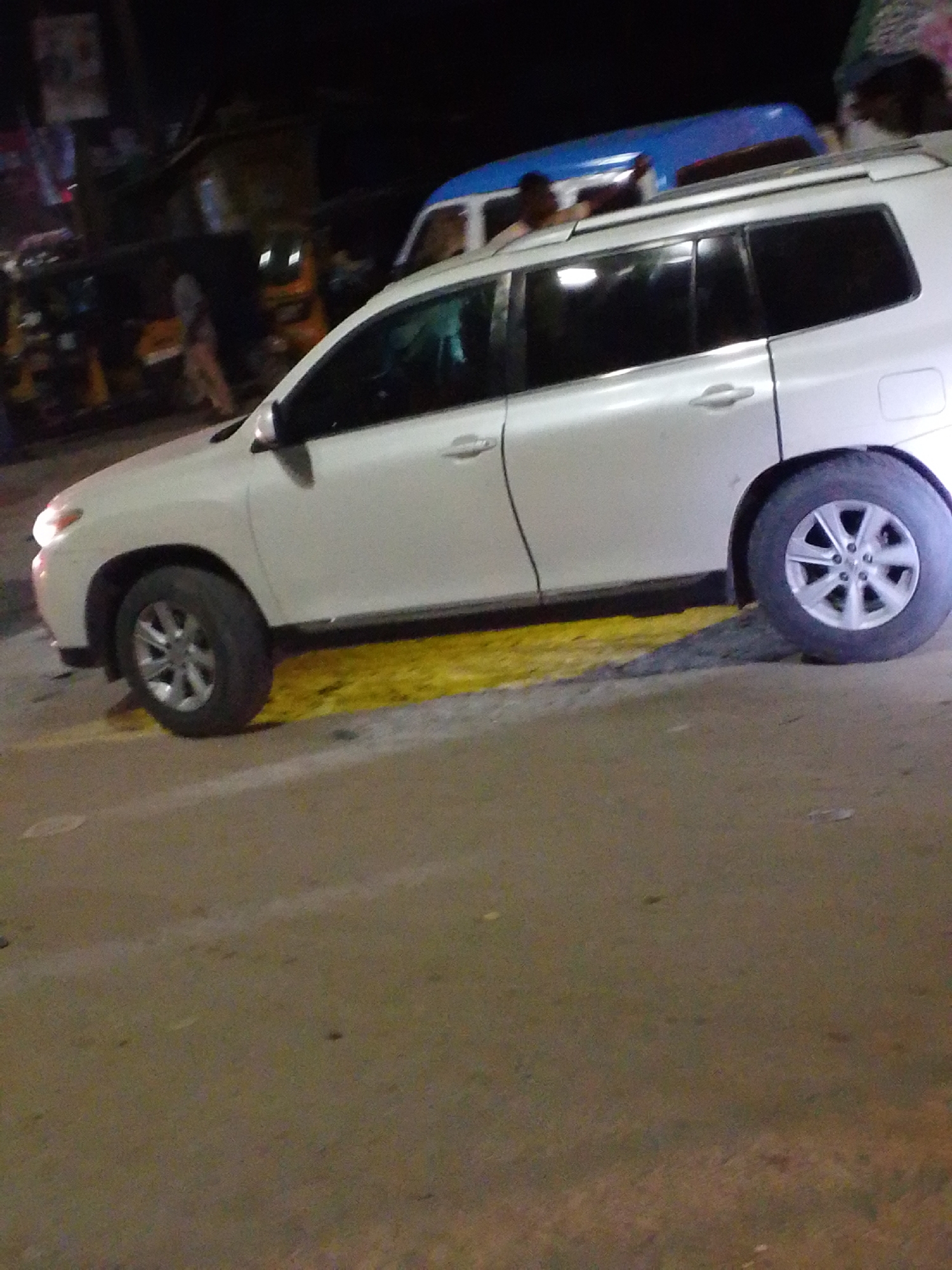 The image shows a white Toyota Highlander SUV parked on a street in Lagos, Nigeria at night. The SUV is parked on a section of yellow painted pavement. Other vehicles, including a blue and white bus and yellow tuk-tuks, are visible in the background, along with people walking on the sidewalk. The lighting conditions indicate it is nighttime.