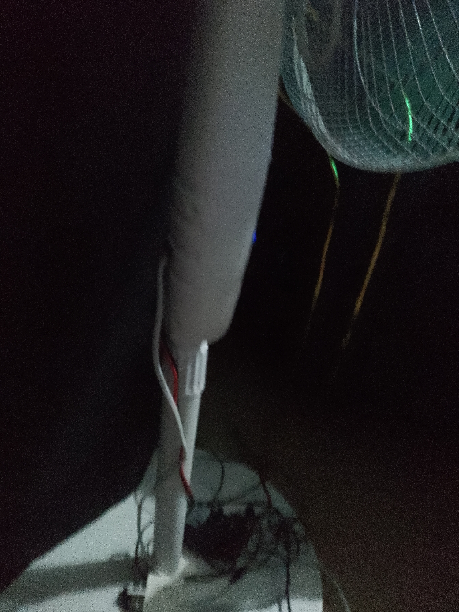 An indoor, eye-level, medium shot captures a close-up of a white stand fan, its cage partially obscured, with a green light visible within. Two yellow cords hang vertically behind the fan. The fan is mounted on a white base, and a section of white tube with red and white wires connecting to a white plastic connector is visible on the fan's pole. The background is dark and out of focus.