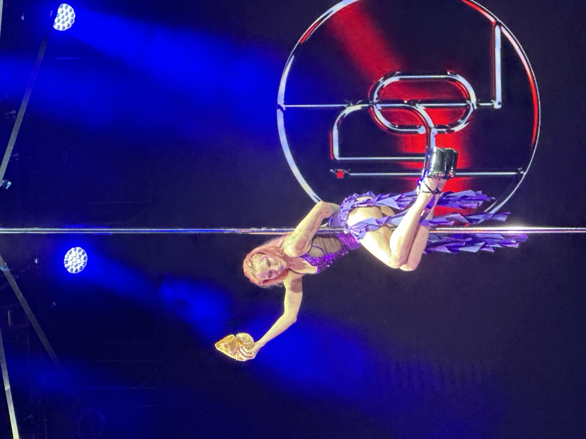 An aerial performer is captured mid-act on a brightly lit stage in Krasnoyarsk, Russia. The central subject is a woman with light reddish hair, dressed in a sparkling purple costume with ruffled, flowing fabric and black high-heeled boots. She is inverted, hanging from a horizontal bar with one leg hooked over it, and extends one arm downwards, holding a golden, textured object that resembles a mask or an ornate piece of art. A gentle smile plays on her face, conveying focus and engagement in her performance.
The background is dominated by intense blue stage lighting, with distinct beams emanating from two visible LED spotlights. Behind the performer, a large, prominent circular structure acts as a backdrop. This structure features metallic, stylized tubing forming what appears to be a bold letter "D" or a unique logo, which is illuminated with red light, creating a strong contrast against the blue stage lights. The setting suggests an indoor evening event or concert, emphasizing the dramatic and dynamic nature of the aerial act. The overall atmosphere is vibrant and theatrical, highlighting the performer's skill and the elaborate stage production.
