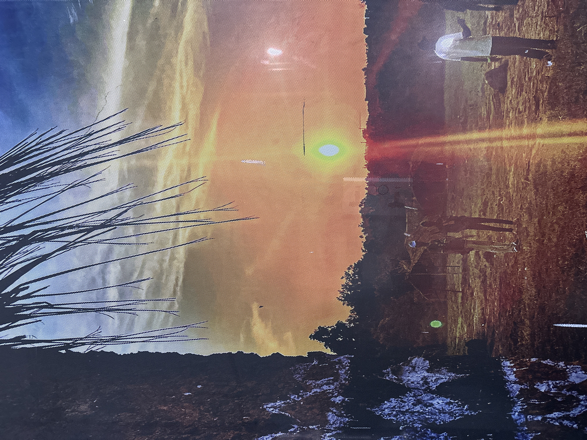 The image depicts an outdoor scene with a sunset or sunrise dominating the sky, transitioning from blue to orange hues. A silhouetted tree is visible on the left. The foreground features a dark landscape, possibly a rocky or uneven terrain with patches of white or light-colored elements, possibly snow or light-colored rocks.
In the middle ground, figures of people are seen standing on a grassy field near buildings, possibly engaging in unspecified activities. The image displays lens flare effects and distortions. The location, Ikeja, Nigeria, is not visually confirmed within the image and is provided externally.