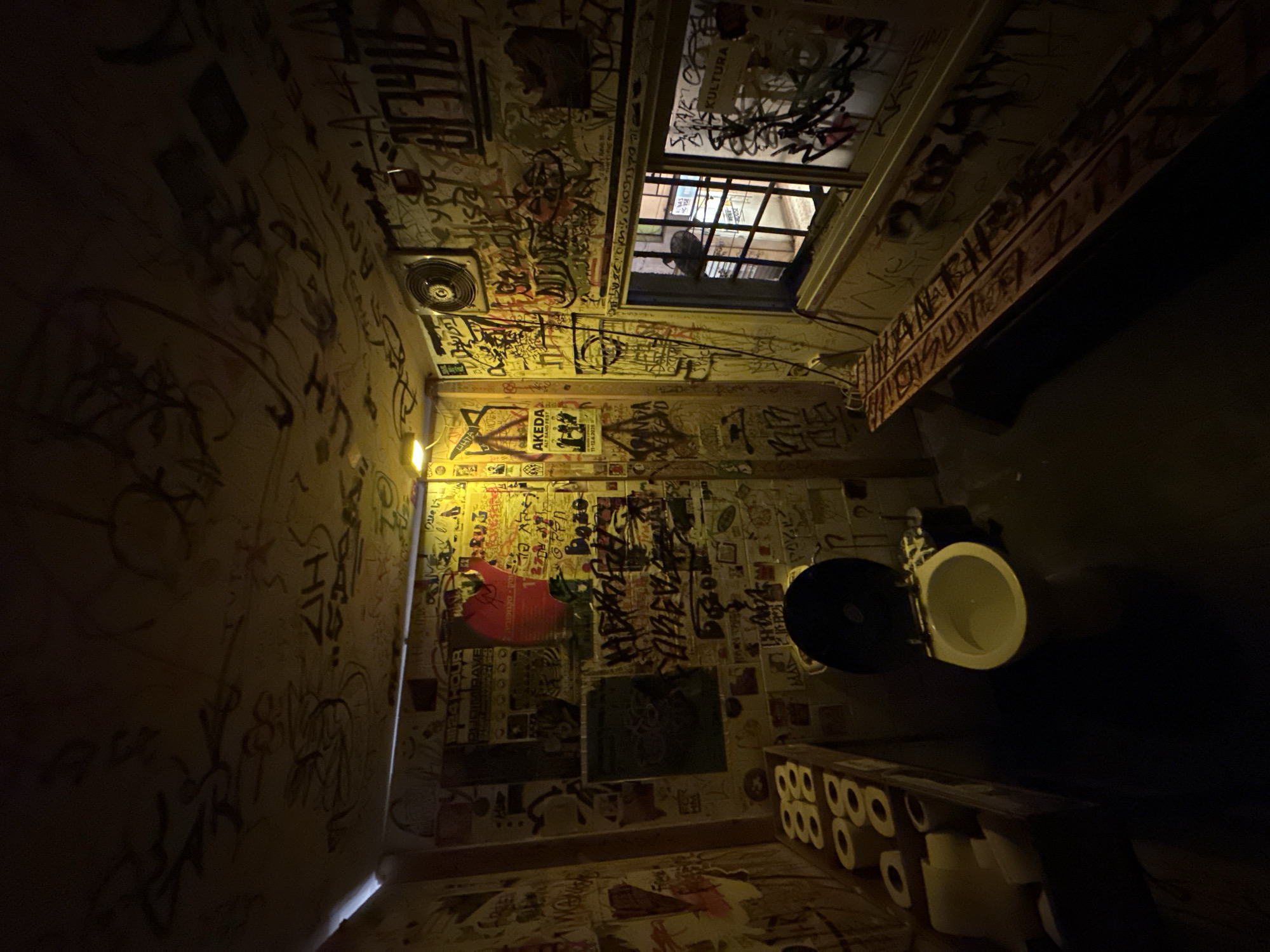This image captures a small, dimly lit room, likely a public restroom, characterized by its heavily graffitied and stickered surfaces. The walls, ceiling, and even parts of the door frame are densely covered with layers of tags, stencils, posters, and stickers, creating a chaotic yet expressive visual environment. The dominant colors are muted yellows and browns from the artificial lighting, contrasting with the dark outlines of the graffiti.
In the upper right corner, a small, gridded window looks out onto a dark exterior, possibly an alley or another building, with limited visibility. Below the window, a white toilet with a dark seat is visible, positioned in a corner. A stack of toilet paper rolls is neatly arranged on a dark surface next to the toilet. A circular ventilation fan is mounted on the ceiling. The room is illuminated by a warm, yellow light source, casting long shadows and highlighting the textures of the wall coverings.
Visible text and details from the graffiti and posters include: "KULTURA" near the window, "AKEDA ALT BAND FEST 11-12.6.2026" on a prominent poster, and various other words and tags such as "UHTA," "JRIMIRAVE," "RUG COMEVA," "NDA," "WEEKEND," and "Misa." The general atmosphere is urban, raw, and full of informal artistic expression, typical of a public space in a vibrant city. No people are visible.