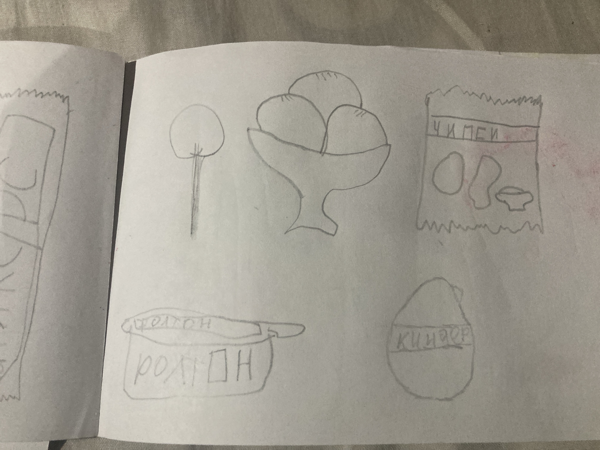The image is a close-up, overhead shot of a sketchbook containing several pencil drawings. The drawings appear to be of food items and a lollipop. In the upper portion of the sketchbook, a drawing of a rectangular object with curved lines and lettering is visible. Below this, a drawing of a hat with the letters "ПОЛ" and "НОП" is depicted. To the right of the hat drawing is a lollipop. In the lower left quadrant, a circular drawing with vertical lettering is present. To the right of this, a drawing of a snack bag with shapes inside and vertical lettering is visible. The drawings are done in pencil on white paper. The image does not contain any recognizable landmarks or contextual information related to the city of Besedy, Russia.