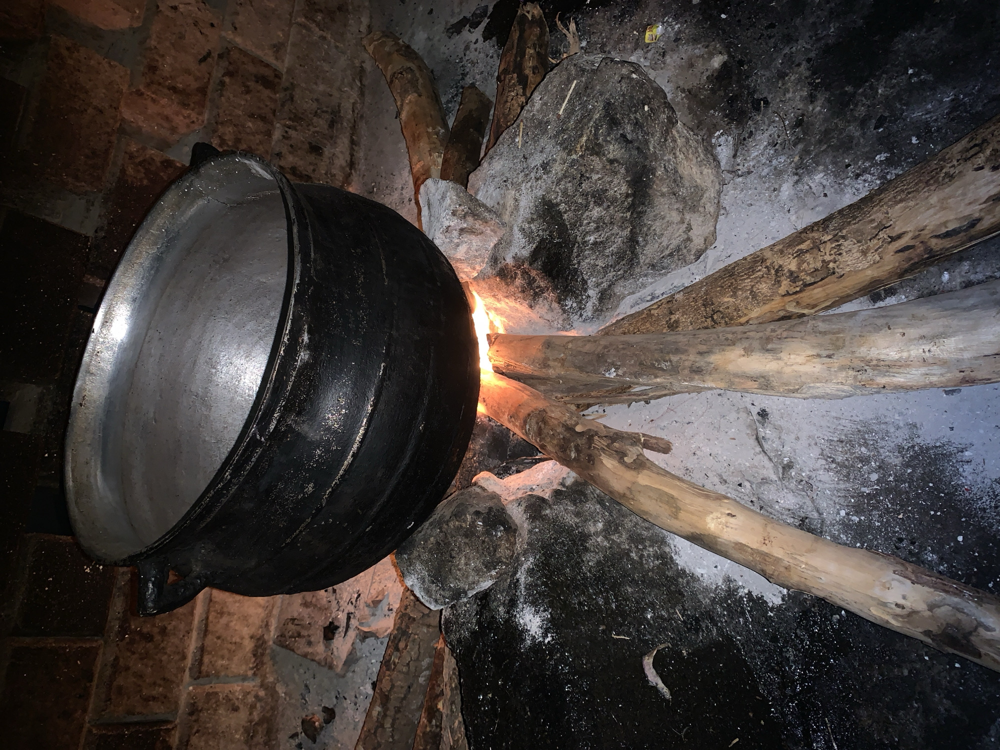 A close-up, low-angle shot shows a large, dark, cast-iron pot suspended over a fire. The pot is black and appears to be old and well-used, with two handles on either side. The fire beneath it is comprised of several logs, with bright orange flames licking upwards, illuminating the underside of the pot and the surrounding stones. The stones are grey and rough, some appear to be cracked or weathered. The ground around the fire pit is dark and appears to be made of earth or ash, with some white powdery residue scattered across it. In the background, a brick wall is visible, constructed from reddish-brown bricks. The overall impression is one of rustic cooking, likely outdoors or in a traditional kitchen setting.