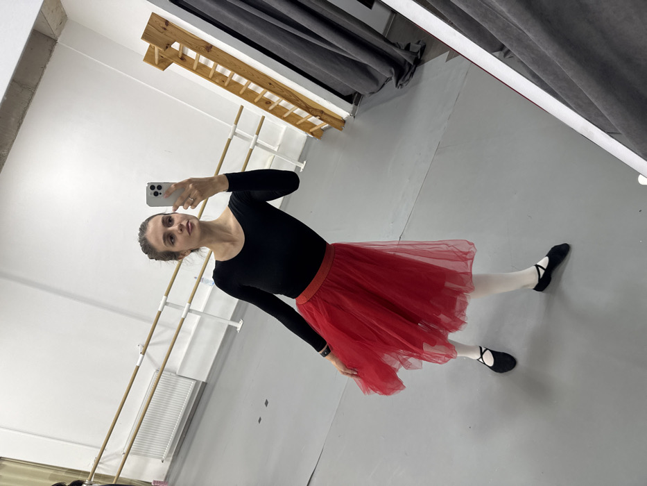A female individual is depicted in a dance studio, location Unknown, Unknown. She is observed taking a mirror selfie using a white smartphone held in her left hand, which displays a ring on her ring finger. Her head is slightly turned to her right. She is wearing a long-sleeved black leotard, a red tiered tulle skirt with a red shimmering waistband, white tights, and black ballet slippers with crisscross straps. A dark band is visible on her right wrist. The studio features light grey flooring and white walls. Two wooden ballet barres with white supports are mounted on the left wall, above a white radiator. To the right, a wooden ladder-like structure with horizontal rungs is visible, along with dark grey curtains partially reflected in the mirror. The scene is well-lit.