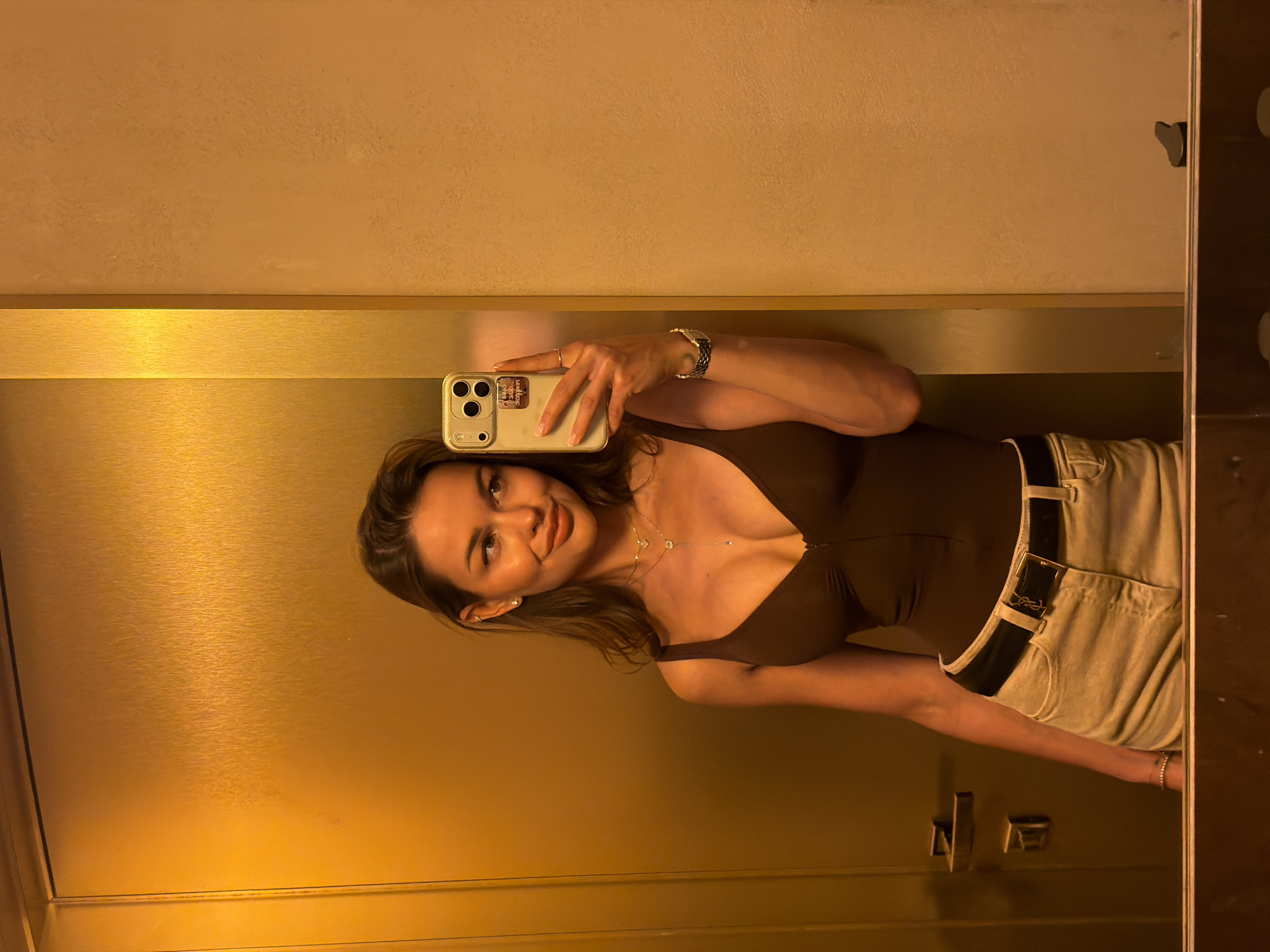In a dimly lit bathroom, a young woman with light brown hair styled in loose waves takes a mirror selfie. She is wearing a dark brown, low-cut leotard with a zipper down the front, and light-colored, high-waisted shorts with a black belt. Her gaze is directed at her reflection, and she has a slight smile on her face. She is adorned with two delicate necklaces, small stud earrings, and a ring on her left hand. A watch with a dark band is visible on her right wrist. The background appears to be a metallic gold wall and a textured beige wall, suggesting a modern and possibly luxurious setting. The overall impression is one of casual elegance and confidence. The scene does not provide any information about the city of Saint Petersburg, Russia.