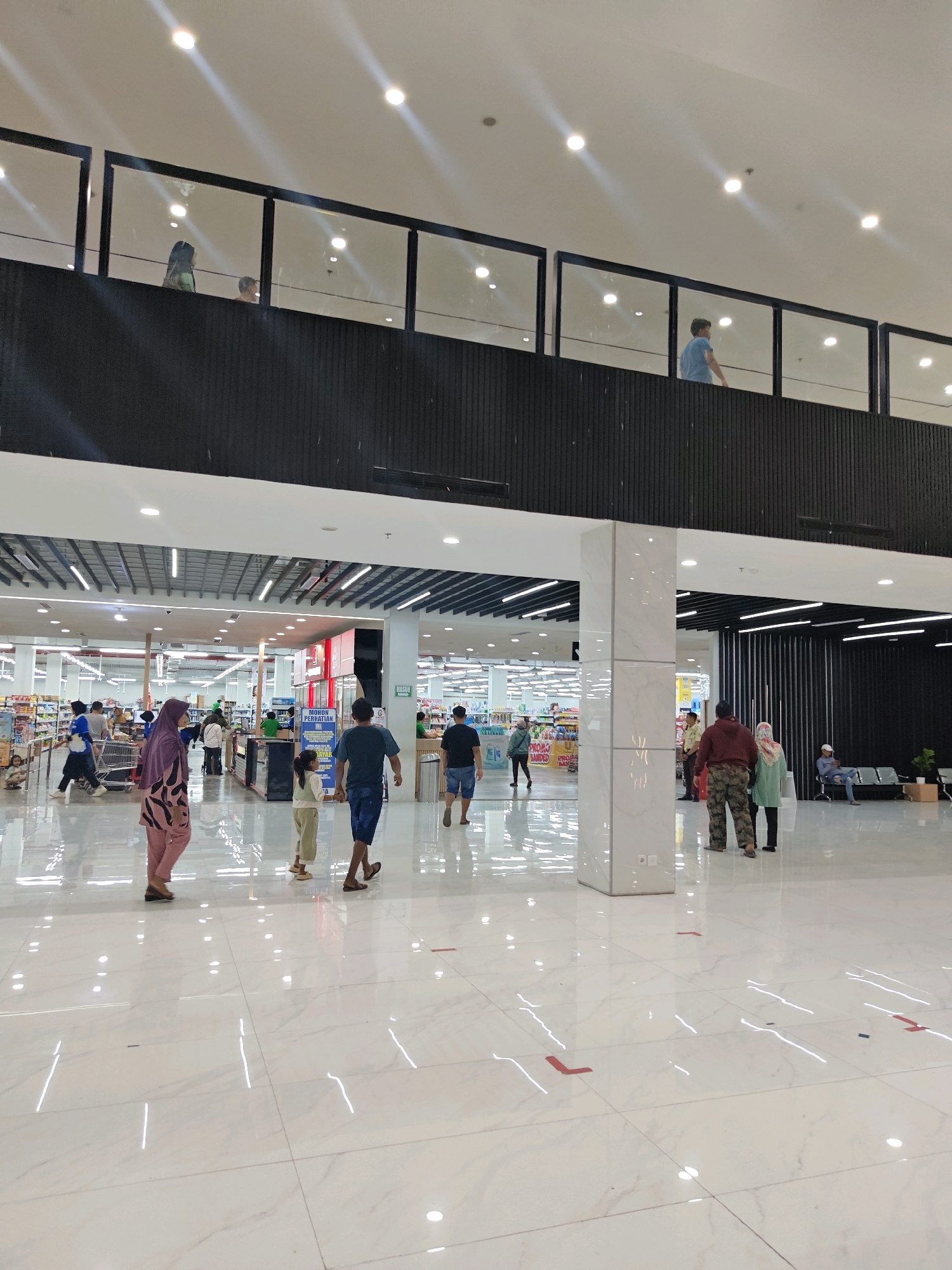 This is an eye-level, full-shot photograph taken inside a modern, multi-level retail environment, likely a supermarket or department store, in Situbondo, Indonesia. The scene is brightly lit by numerous overhead fluorescent lights and illuminated ceiling panels. The floor is a polished white marble with faint grey veining, reflecting the lights and the activity above and around.
In the foreground, a man in a dark t-shirt and blue shorts walks away from the camera, his right hand holding that of a young girl in a light-colored top and pants. To their left, a woman in a pink headscarf and patterned pink pants walks away from the camera. Further left, a man in a blue t-shirt and dark shorts walks away from the camera, pushing a shopping cart.
In the middle ground, on the main level, several individuals are seen browsing merchandise shelves. One woman in a light-colored hijab and pink patterned outfit is visible, as is a man in a dark blue shirt and light-colored pants. A cashier station with a sign that reads "MOHON PERHATIAN" (Please Pay Attention) is visible, with people interacting with staff. Other shoppers are scattered throughout the aisles.
In the background, the store extends with rows of shelves stocked with various products, some with prominent "PROMO LANDED" signage.
Above, a second level is visible, separated by a black horizontal barrier and framed by sleek black vertical supports. A glass railing allows views onto this upper level, where at least four individuals are present. One person in a green top and dark hair is looking down, another in a blue t-shirt is looking to the side, and two others are visible further back, one in a blue top and another in a light-colored shirt.
To the far right of the middle ground, a seating area with rows of chairs is present. One person in a light-colored shirt is seated, and another person in a light blue hijab is standing nearby.
The overall impression is a busy, well-maintained retail space with shoppers engaged in everyday activities. The lighting is uniform and bright, creating a clean and modern aesthetic.