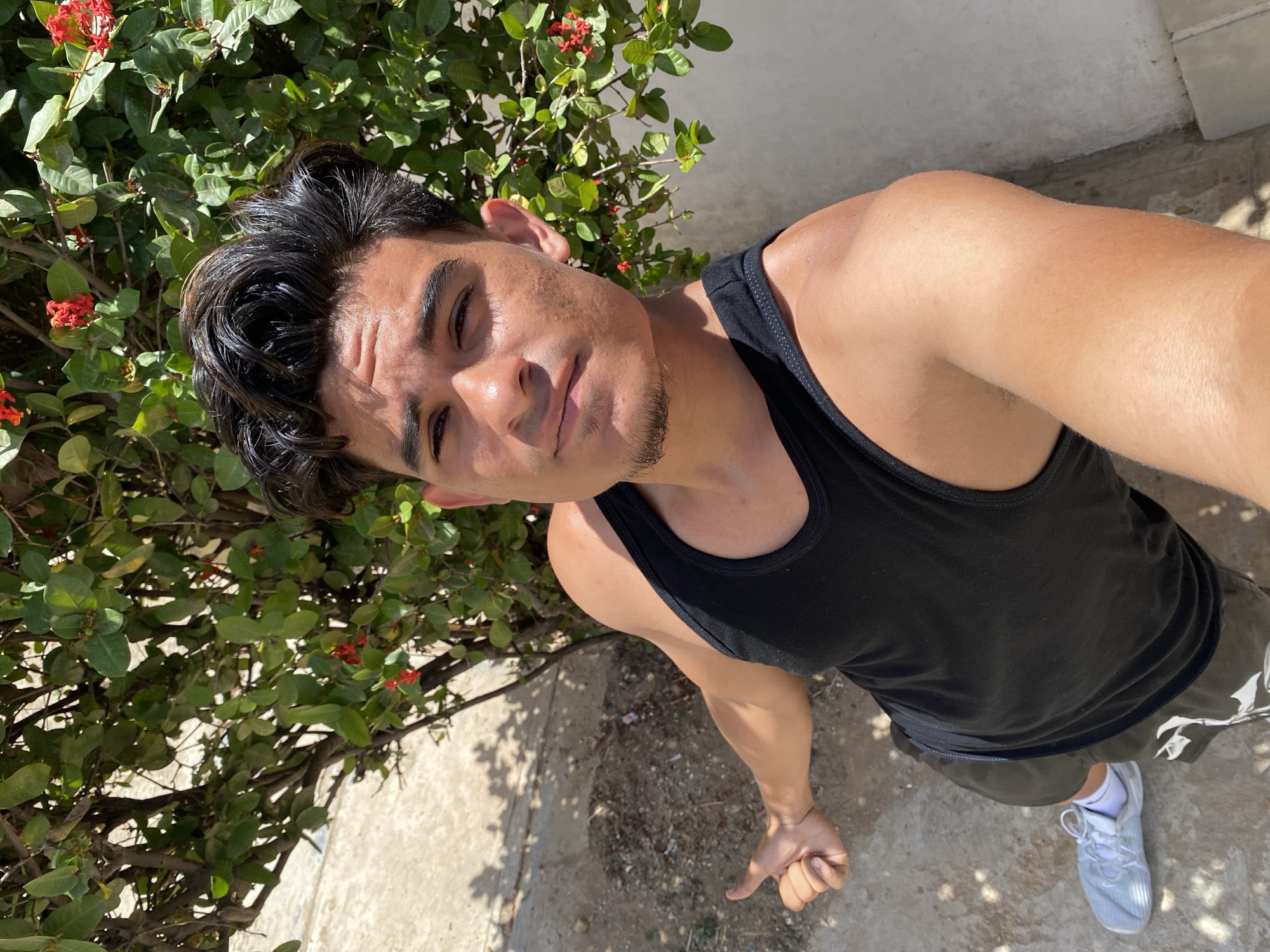 A medium-shot, high-angle photograph shows a light-skinned, dark-haired male wearing a black tank top and dark shorts in an outdoor setting. The man is positioned near a flowering bush with green leaves and small red blossoms. The ground is concrete with scattered debris. The location is identified as Juan Griego, Venezuela. The man is looking up at the camera. He is wearing a white sneaker on one foot, and his hand is clenched near the bottom edge of the frame.