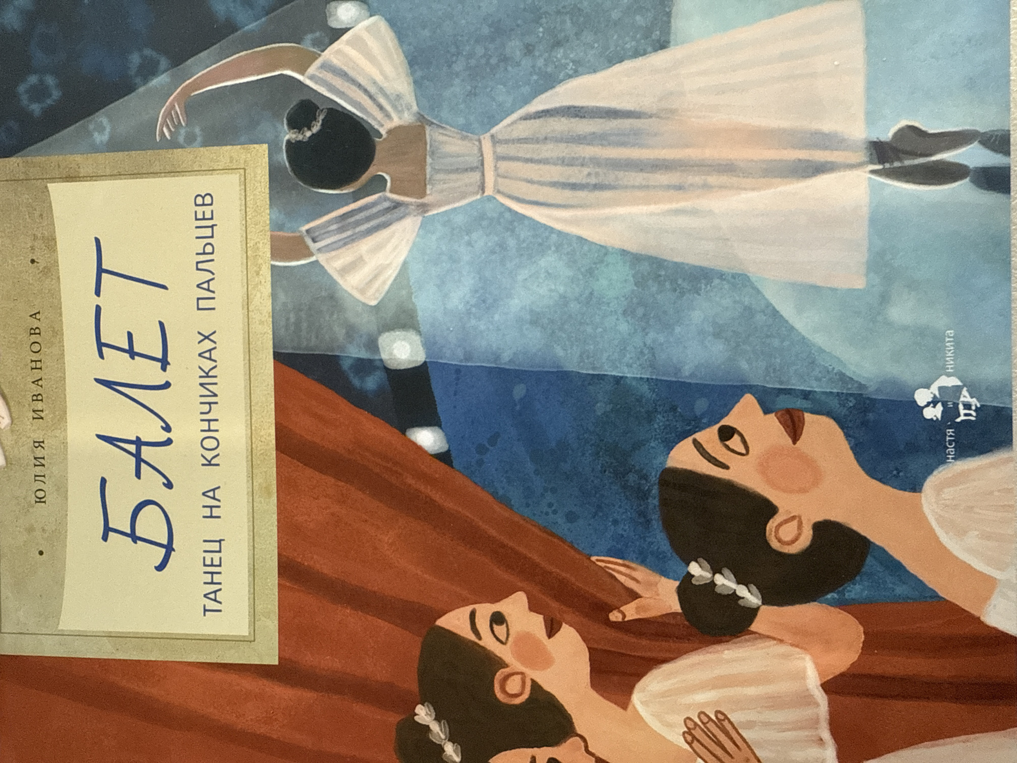 The image is a book cover. It depicts a ballet scene with three ballerinas in white dresses. One ballerina is positioned in the upper part of the image, leaping in the air. Two ballerinas are lying on what appears to be a stage draped with red fabric. The background is a mottled blue color.
The cover has text in the Russian language: "Юлия Иванова" (Julia Ivanova) and "БАЛЕТ" (Ballet), with the subtitle "ТАНЕЦ НА КОНЧИКАХ ПАЛЬЦЕВ" (Dance on the Tips of Toes). There is also a logo with the word "настя" (Nastya) next to it.
The location, Votkinsk, Russia, is not explicitly stated in the image but would be inferred from context. The image is likely from a book published in Russian, potentially by an author from or about the region, but this cannot be definitively determined from the image alone.