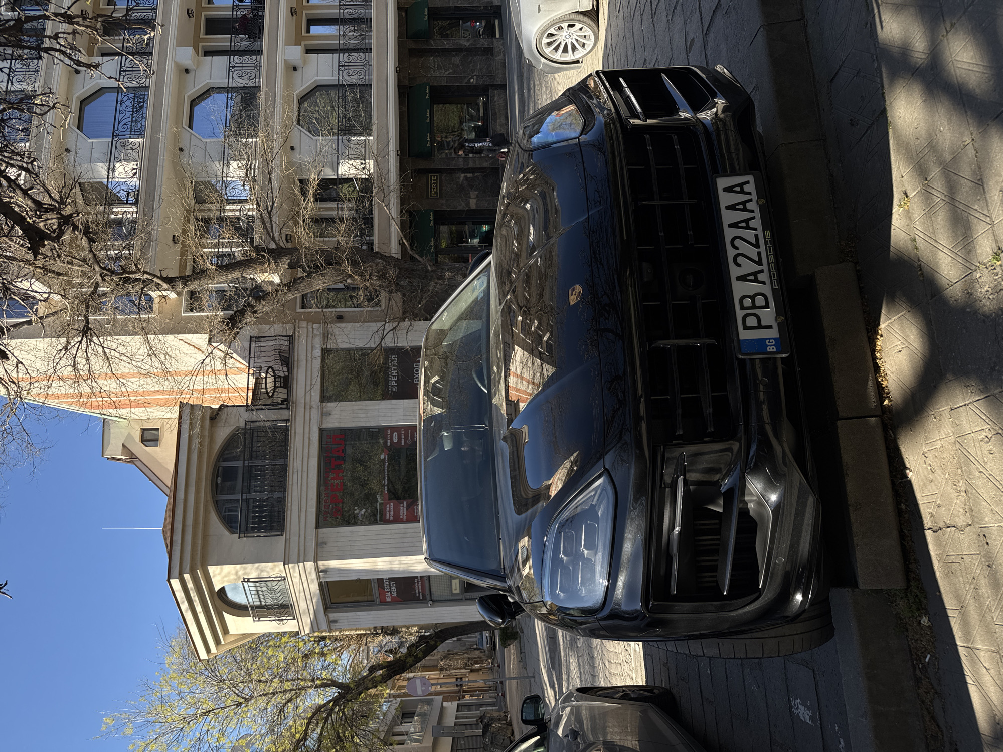 A black Porsche SUV is parked on a cobblestone street in Plovdiv, Bulgaria. The license plate reads "PB A22 AAA". The front of the car faces the viewer. Behind the car is a multi-story building with arched windows and wrought iron detailing. A tree with bare branches is visible in front of the building. Sunlight casts shadows on the street. The weather appears clear.