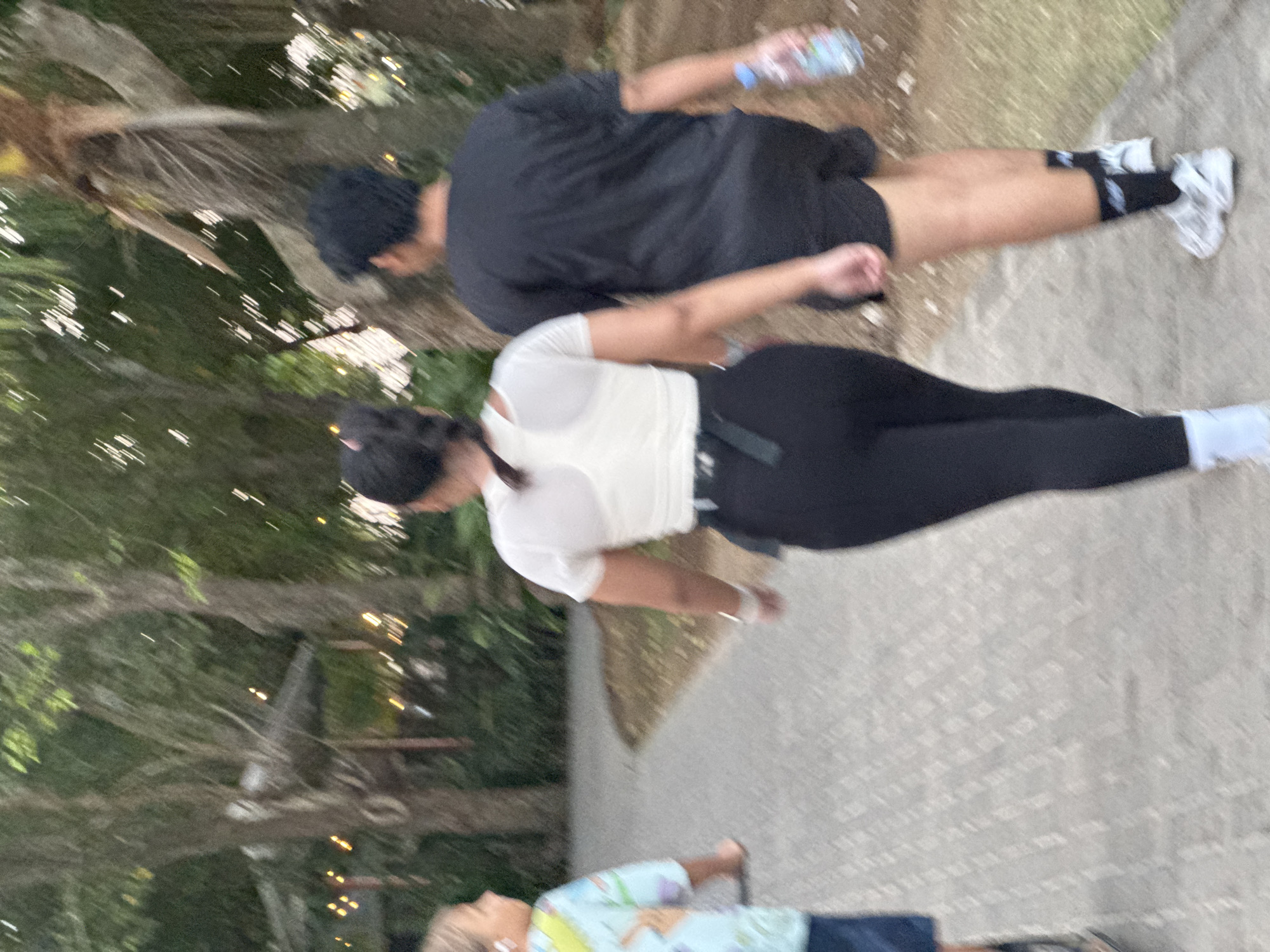 In the city of Kuta Selatan, Indonesia, three individuals are captured walking along a paved pathway. The person on the left, a child, is wearing a light blue shirt with a cartoon print and dark shorts. They are seen from the side, with their left hand raised. In the center, a person with dark hair pulled back in a braid is wearing a white athletic top and black leggings. They are walking away from the camera, with their left arm slightly extended. On the right, a person with dark hair and wearing a black t-shirt and black shorts is also walking away from the camera. They are holding a clear plastic water bottle in their right hand. The environment is lush with trees and foliage, suggesting a park or a tropical outdoor setting. String lights are visible in the background, indicating it may be evening or early morning. The pathway is made of interlocking pavers.