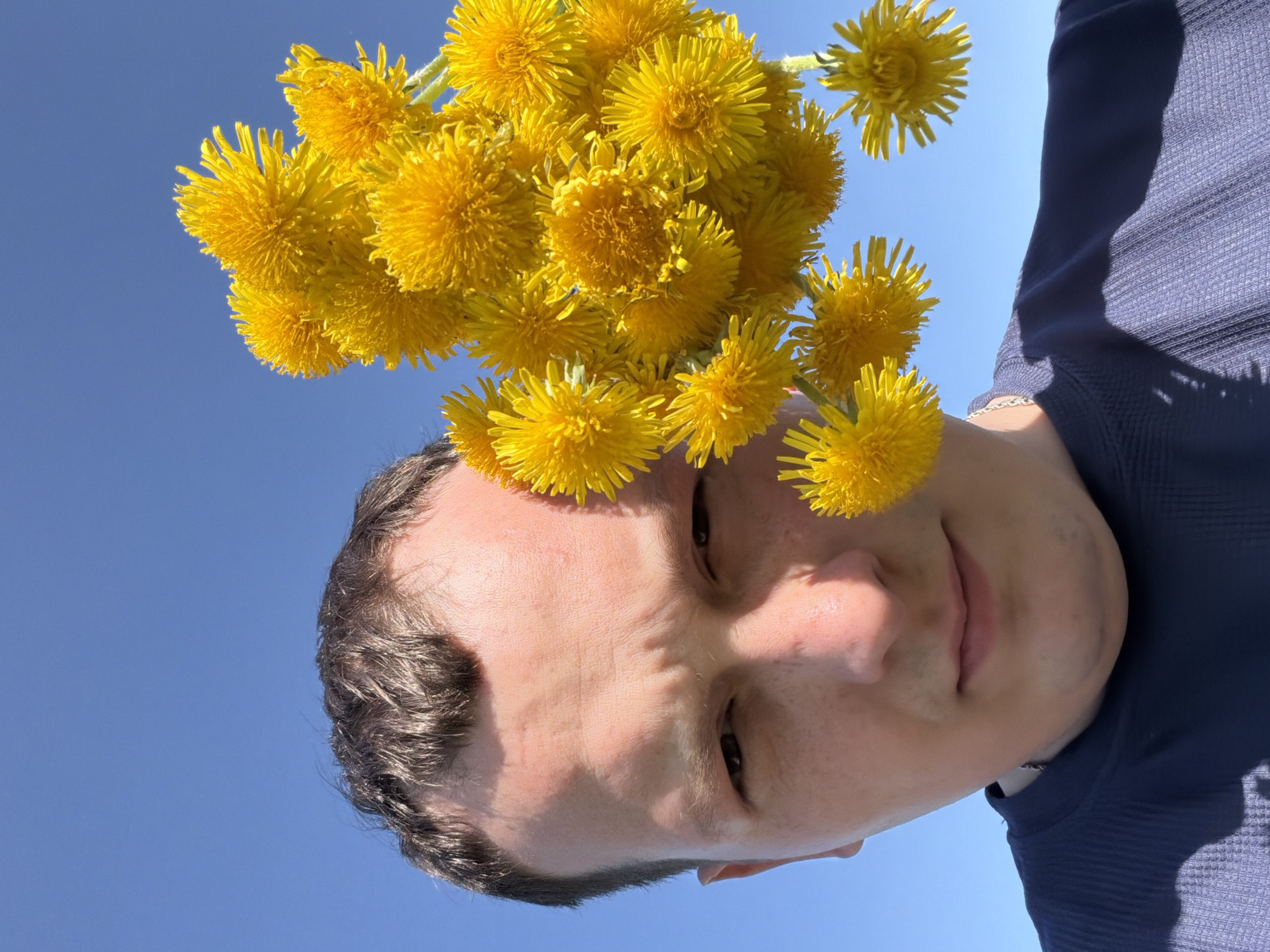 A low-angle shot of a man, possibly in his late 20s or early 30s, holding a bouquet of bright yellow dandelions against a clear blue sky. The man's face is prominently featured, with short dark hair, light skin, and dark eyes. He is wearing a dark blue long-sleeved shirt. The image appears to be taken outdoors, possibly in the city of Кемуль, Russia, as indicated by the instructions. The location is implied by the absence of specific landmarks or distinguishing features. The focus is on the man and the dandelions, with the sky serving as a backdrop.