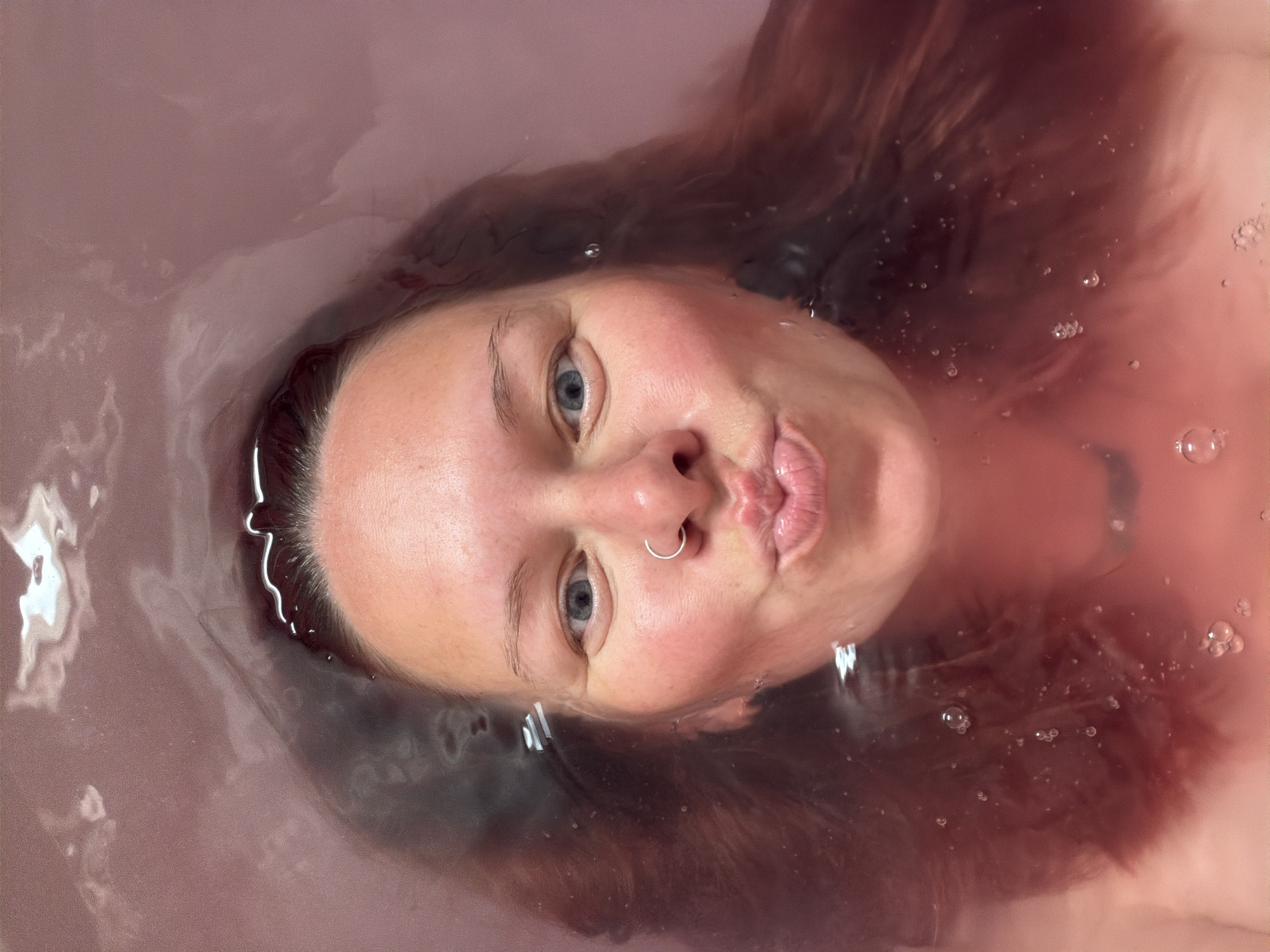 A light-skinned person with blue eyes and a nose ring is partially submerged in water, likely a bathtub. The water has a reddish tint, possibly from bath salts or dye. The person's hair is brown and appears wet. They are looking directly at the camera with puckered lips. The environment is blurred by water. No information indicates that this photo was taken in Chaikovsky, Russia.
