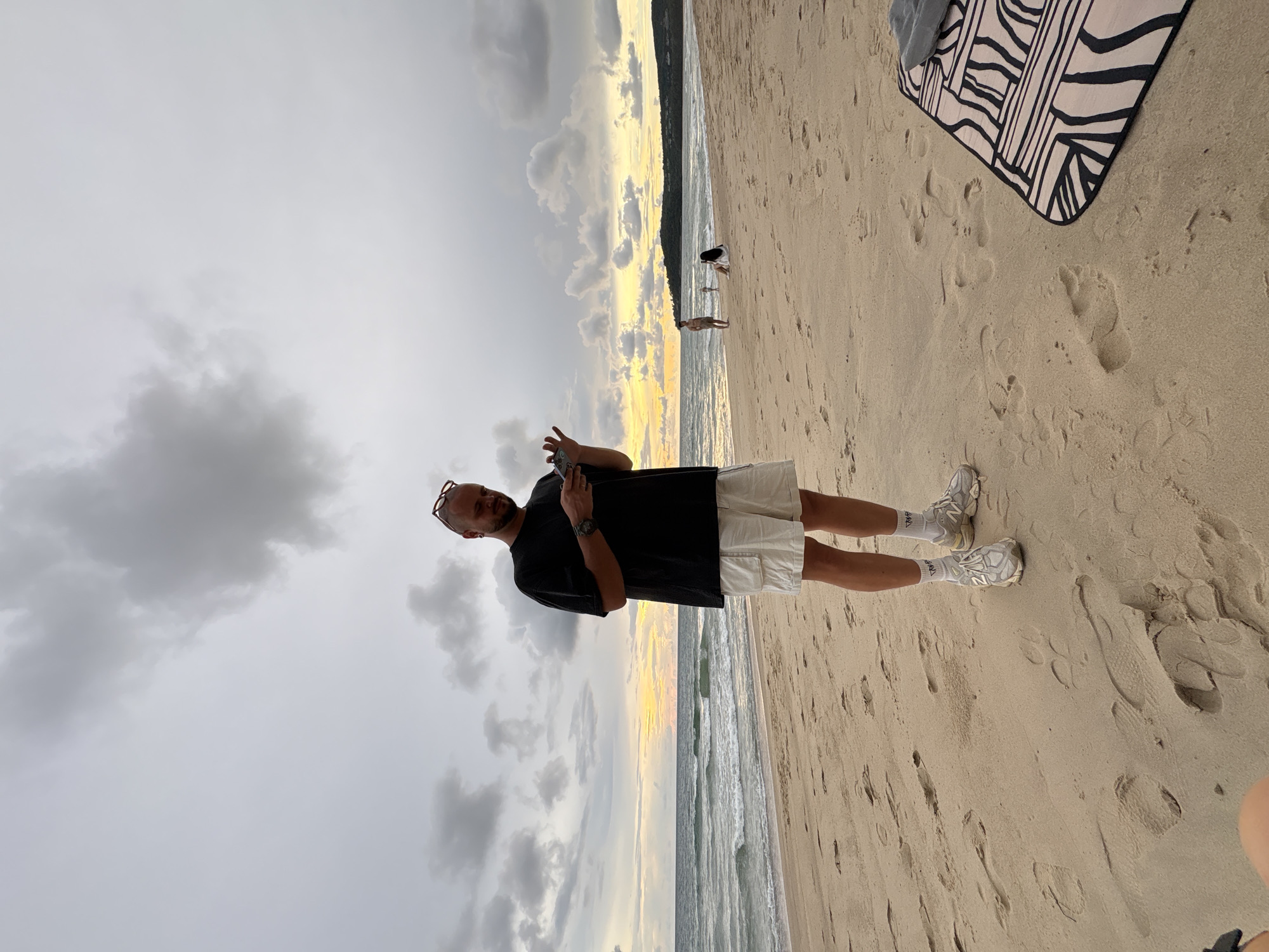 This is a photograph taken at Choeng Thale, Thailand, featuring a man standing on a sandy beach. He is wearing a black t-shirt, white shorts, white socks, and grey sneakers. He has a watch on his left wrist and is holding a mobile phone in his right hand, with his fingers extended as if interacting with it. He is also wearing sunglasses pushed up onto his head.
The scene is a beach with the ocean and waves visible in the background, and the sand is marked with footprints. Further back, a thin strip of land with trees or vegetation can be seen along the coastline. In the distance, other people are visible on the beach, including one person in black and white on the right, and a figure further out by the water.
The sky is overcast with clouds, but there are hints of sunset colors, with streaks of yellow and orange light visible near the horizon, suggesting the time of day is late afternoon or early evening. The overall mood appears relaxed and casual, consistent with a beach setting. A black and white patterned blanket is also visible on the sand in the upper right portion of the image.