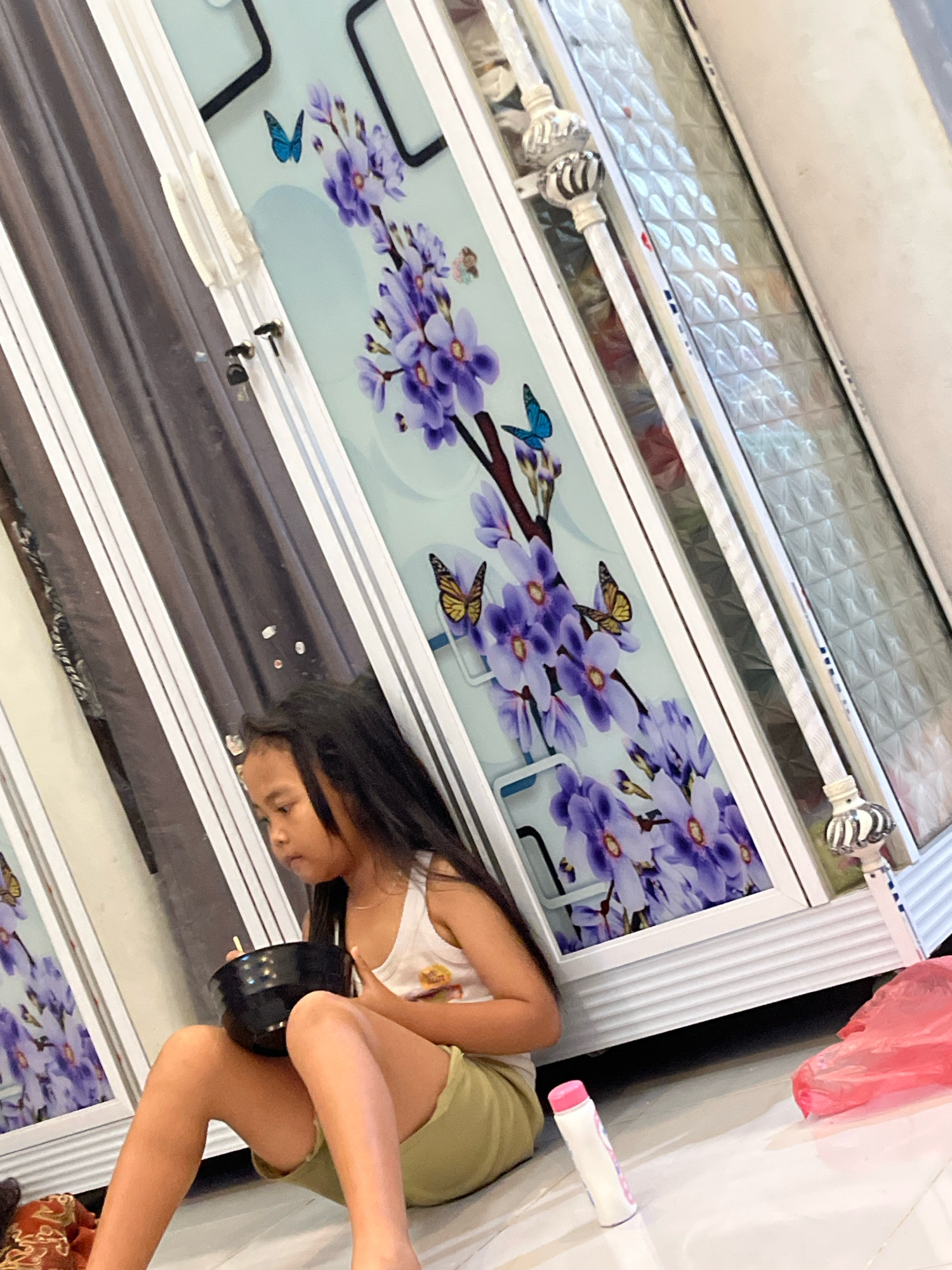 A young Indonesian girl with long, dark hair is sitting on a tiled floor in what appears to be a residential setting in Kebumen, Indonesia. She is wearing a white tank top and olive green shorts. She is holding a black bowl in her lap and appears to be eating or interacting with its contents. To her left are closet doors with decorative panels featuring purple flowers and butterflies. A pink plastic bag and a white bottle with a pink cap are visible on the floor near her. The overall lighting suggests an indoor environment, possibly during the daytime.