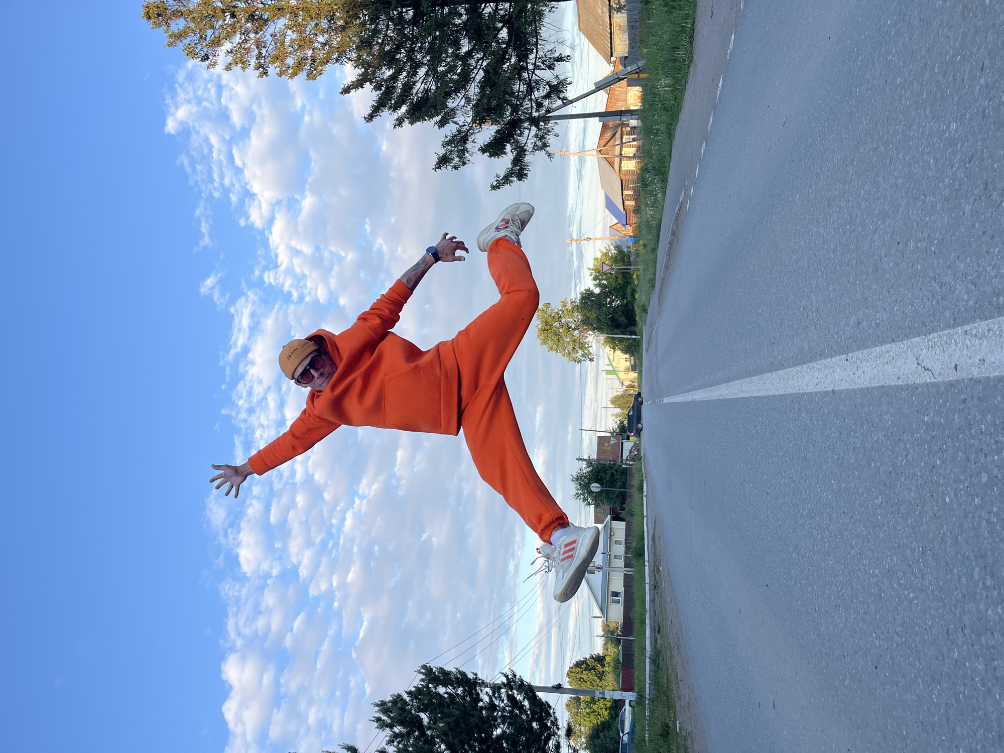 The image captures a person mid-air, performing an energetic pose that resembles a leap or kick, against a bright sky and road background. The primary subject is an individual, likely an adult, dressed in a vibrant, monochromatic orange tracksuit, comprising a hoodie and sweatpants. They wear a light brown beanie with the word "Human" faintly visible, and dark sunglasses. White sneakers with distinctive red stripes, likely "Adidas" given the design, are visible on their feet. A smartwatch is worn on their left wrist, and tattoos are visible on their arms.
The activity depicts a dynamic, suspended moment, suggesting a jump, dance, or playful act. The person has one leg extended backward and upwards, while their arms are spread out to the sides, creating a sense of balance and motion.
The setting is an outdoor scene, likely a suburban or semi-rural area in Chaikovsky, Russia. The sky is a clear blue with scattered white cumulus clouds, indicating fair weather. The ground features a paved road with a solid white line marking its edge, bordered by green grass. In the background, various trees—both coniferous and deciduous—are visible, along with distant houses or buildings and utility poles with wires. The lighting suggests it is either late afternoon or early evening, with soft, ample light. The overall mood conveyed by the pose is one of exuberance and playfulness.