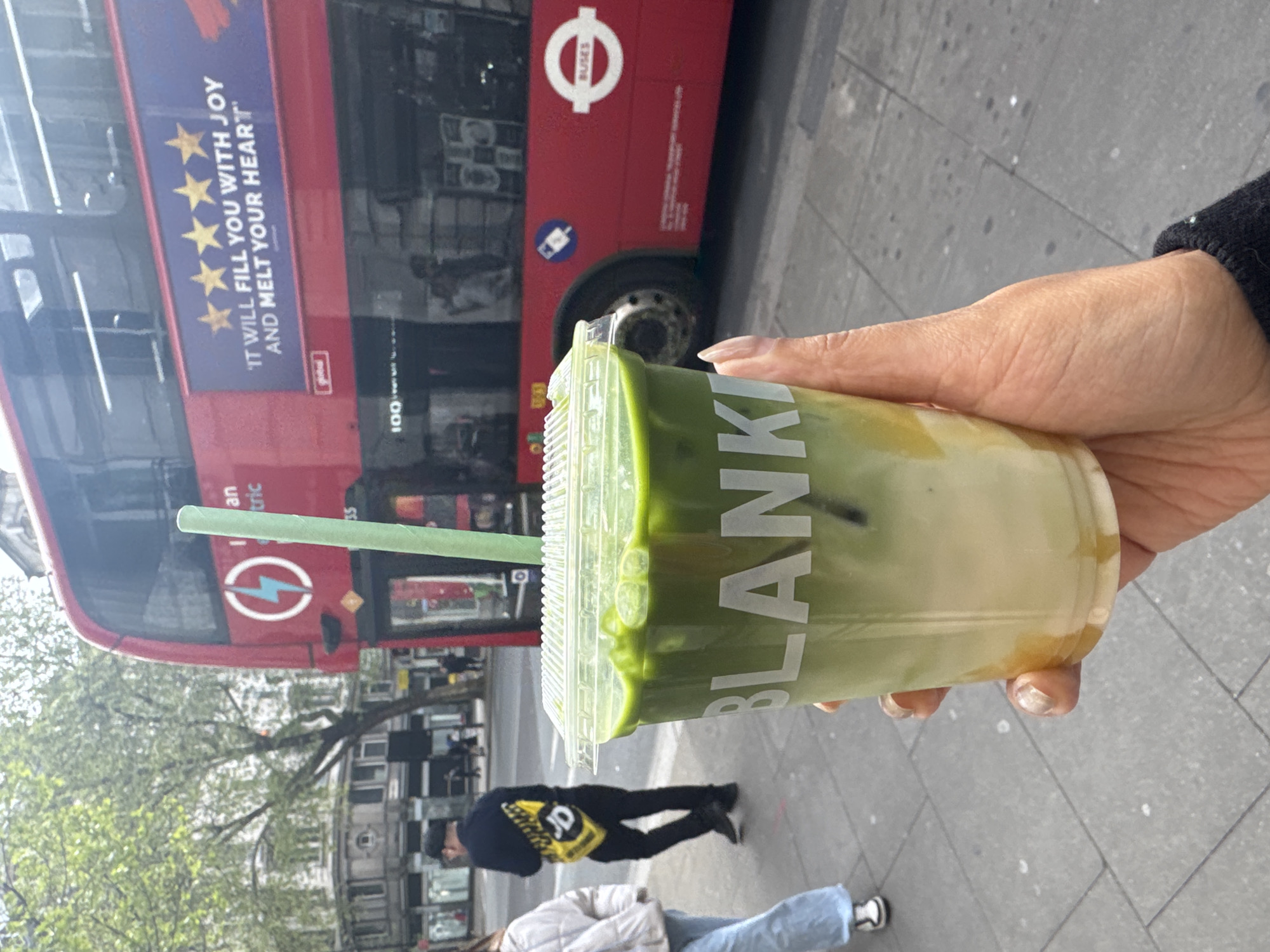 A hand holds a green-tea beverage called "Blanka" in a clear, plastic cup with a green lid and straw. Behind the cup is a red London double-decker bus with the words "IT WILL FILL YOU WITH JOY AND MELT YOUR HEART" beneath five stars. The word "Buses" is also visible on the side of the bus. People are walking in the background on a grey sidewalk.
