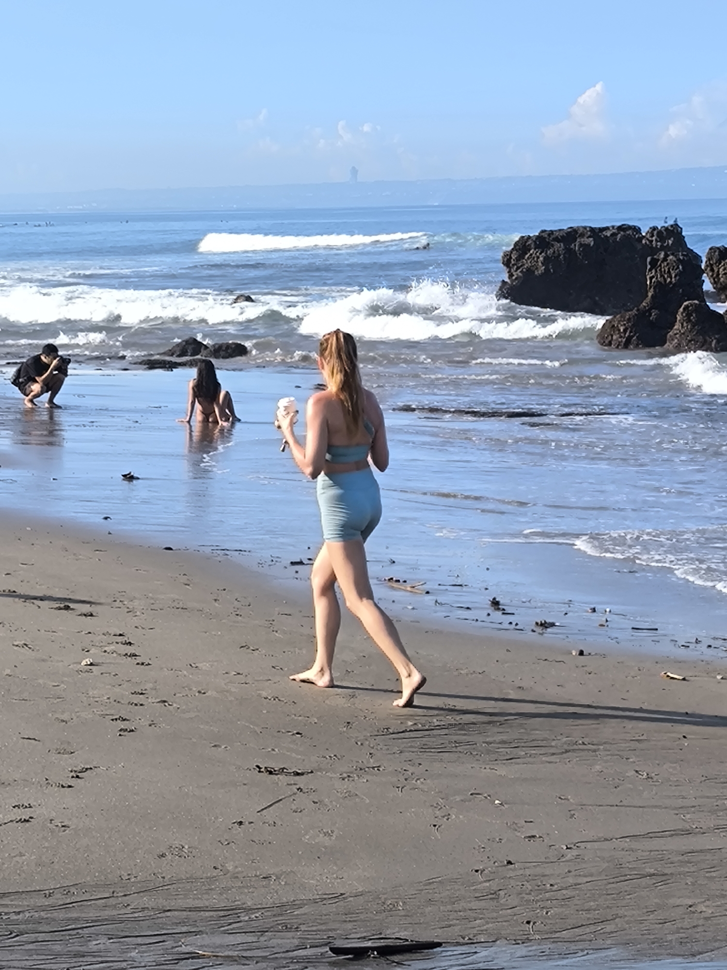 The photograph captures a daytime scene on a beach in Canggu, Indonesia. The environment includes a sandy beach, ocean waves, and rock formations. There are three people visible: a woman in light blue athletic wear walking barefoot toward the right of the frame while holding what appears to be a cup or container; a woman wearing a bikini sitting in the shallow water with her back to the camera; and a person wearing a black shirt kneeling and facing the woman in the bikini, possibly taking a photograph. The sky is light blue with some clouds. Surfers can be seen on the ocean in the distance.