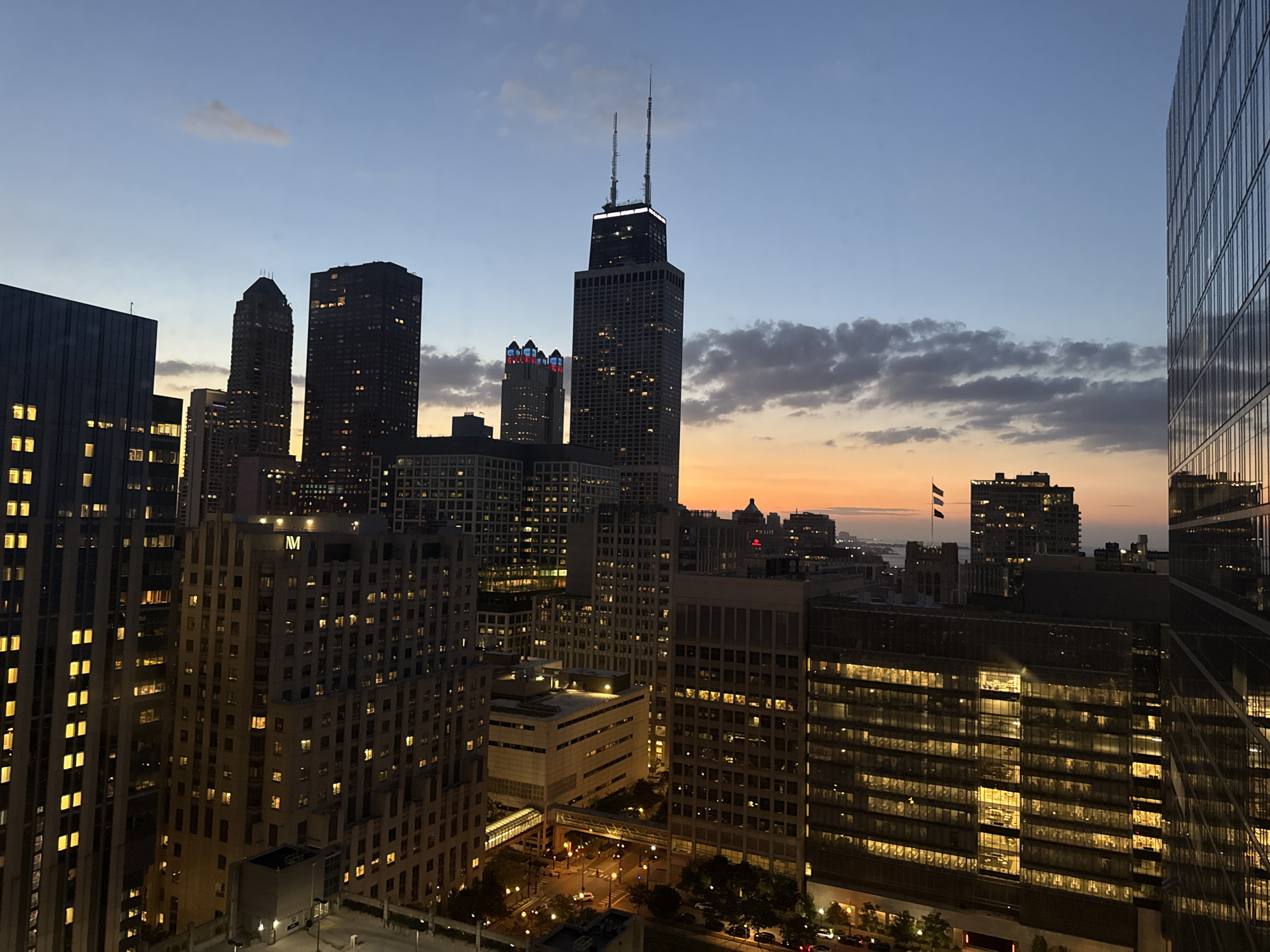 The image captures a dusk scene overlooking the Chicago skyline. Prominent buildings, including the Willis Tower with its distinctive spires, are visible, illuminated by internal lights. Other skyscrapers, some with colored lighting, dot the cityscape. The sky displays a gradient of blue transitioning to orange near the horizon, with scattered clouds. The foreground features the reflective facade of a modern glass building, mirroring the city lights and sky. Below, a street level view reveals illuminated roads, streetlights, and parked vehicles, suggesting ongoing activity as night falls. The overall atmosphere is one of urban dynamism and the transition from day to night.