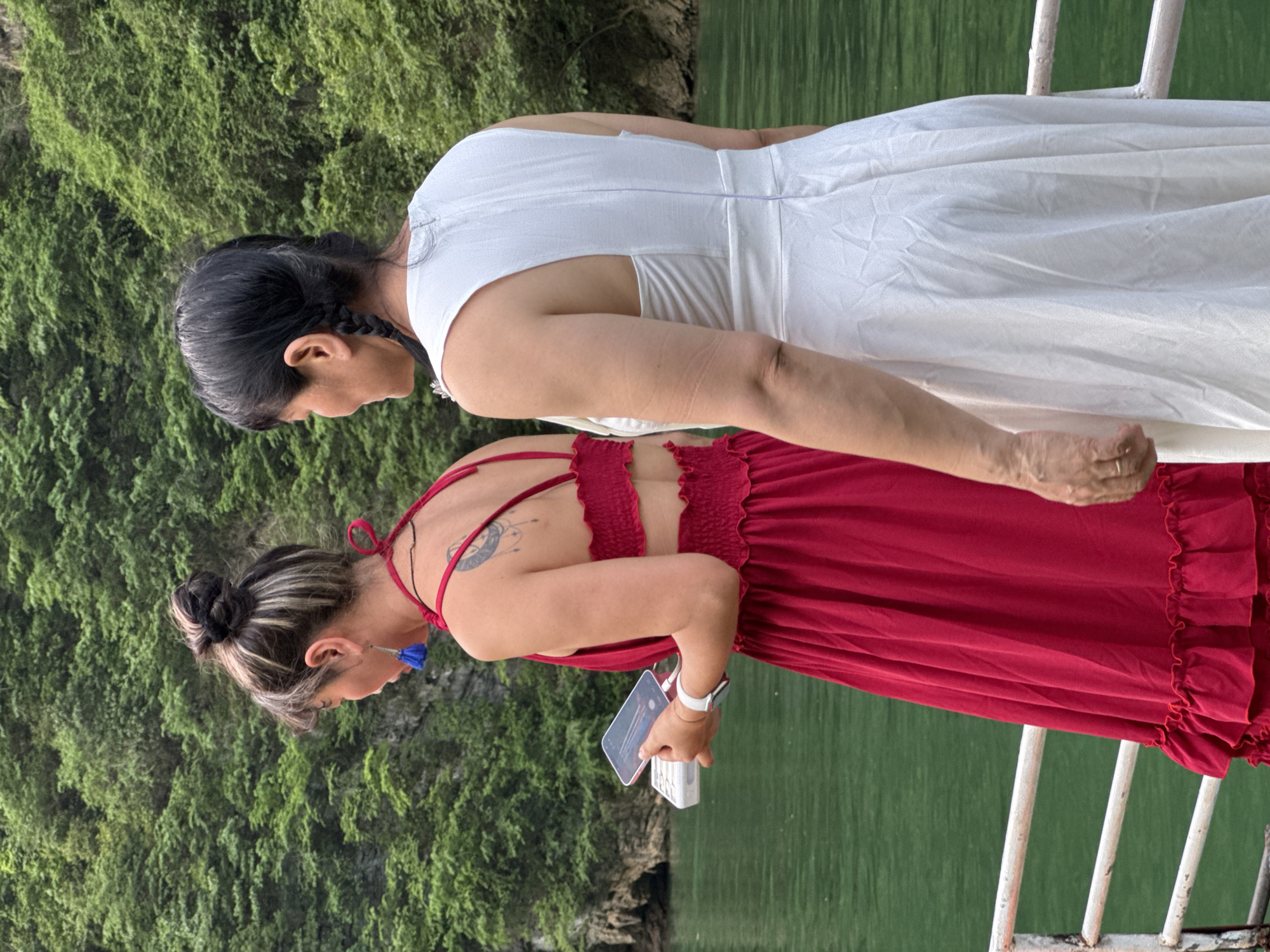 Two women stand next to each other on what appears to be a boat or platform with a white metal railing, against a backdrop of lush green vegetation and water. The woman on top is wearing a white dress. Her hair is black and pulled back. The woman on the bottom is wearing a red dress with a ruched back detail. She holds a smartphone and a small white object in her hands. She is also wearing a white smartwatch and blue tassel earrings, and her hair is pulled up in a bun. The picture was taken in Đồng Văn Commune, Vietnam.